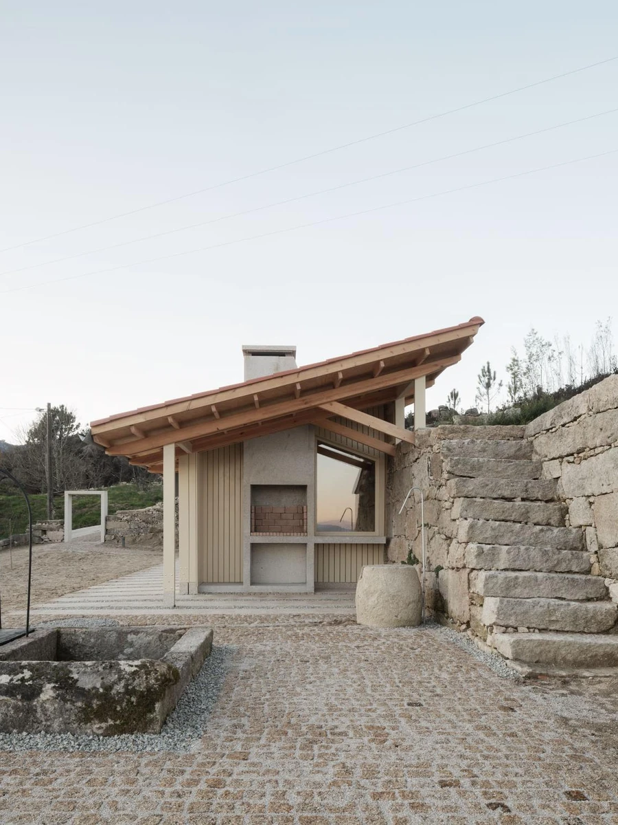 Muimenta Multipurpose Social Center by FIRM + BASED, photographed by Luis Díaz Díaz — A timber pavilion under a tile roof meets a granite terrace wall, with a well and stone trough set into a cobbled courtyard.