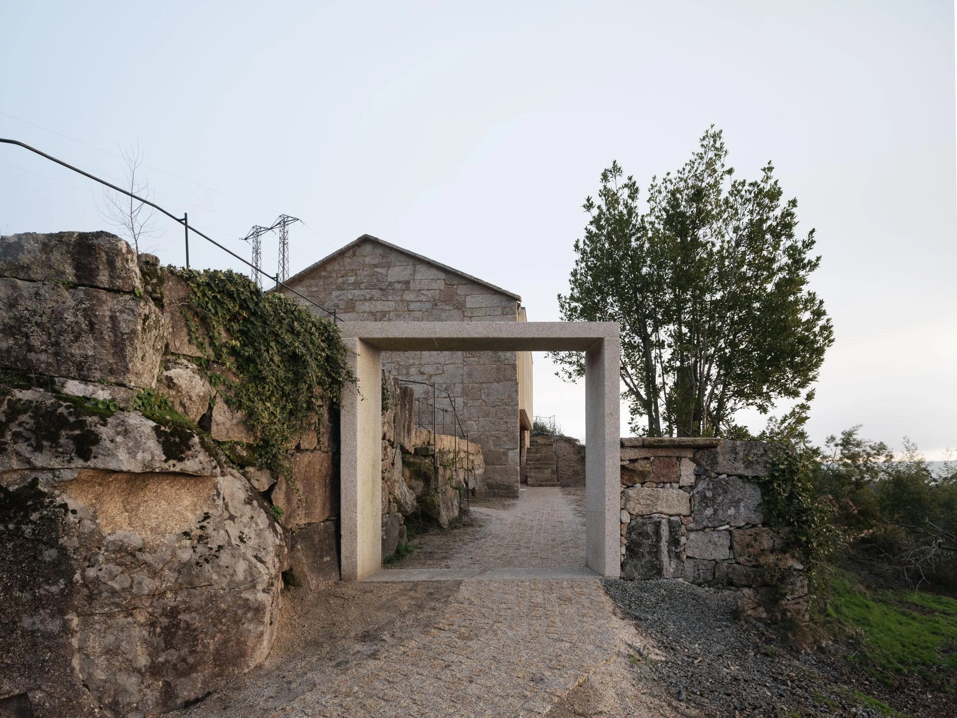Muimenta Multipurpose Social Center by FIRM + BASED, photographed by Luis Díaz Díaz — A granite portal frames a cobbled path between dry-stone terrace walls, leading toward the restored gable of the old house.