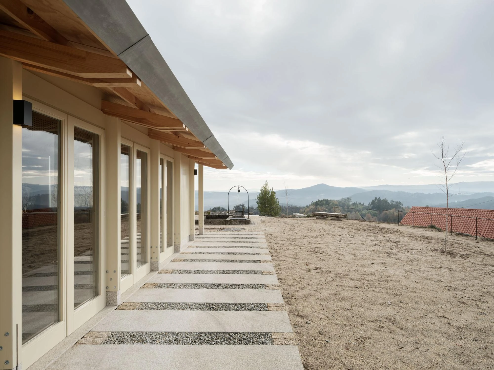 Muimenta Multipurpose Social Center by FIRM + BASED, photographed by Luis Díaz Díaz — A long glazed facade under deep timber eaves opens onto a paved terrace that drops away to the Ourense mountains.