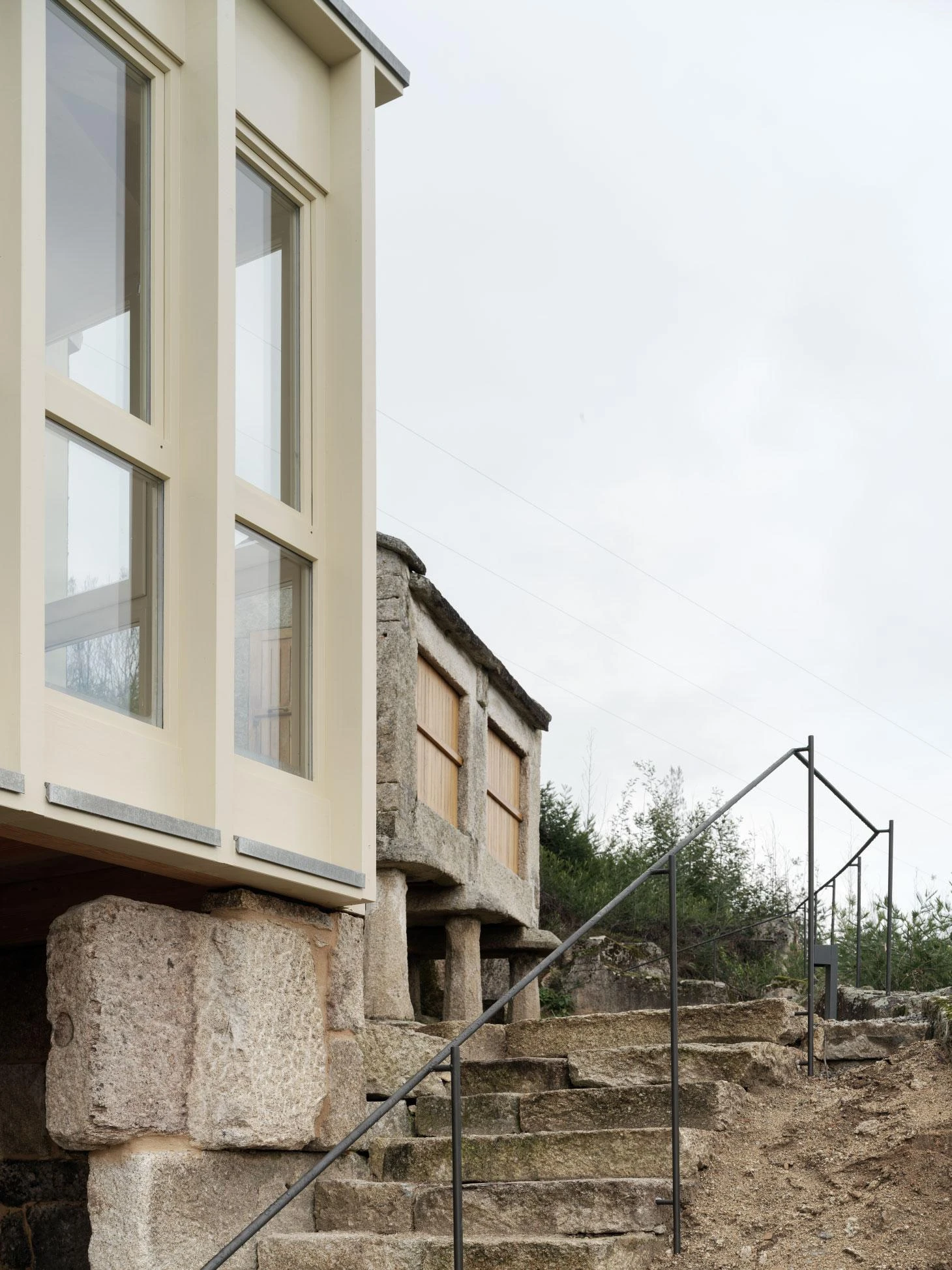 Muimenta Multipurpose Social Center by FIRM + BASED, photographed by Luis Díaz Díaz — A glazed corner window sits on a granite base beside the raised stone horreo, reached by a run of rough granite steps.