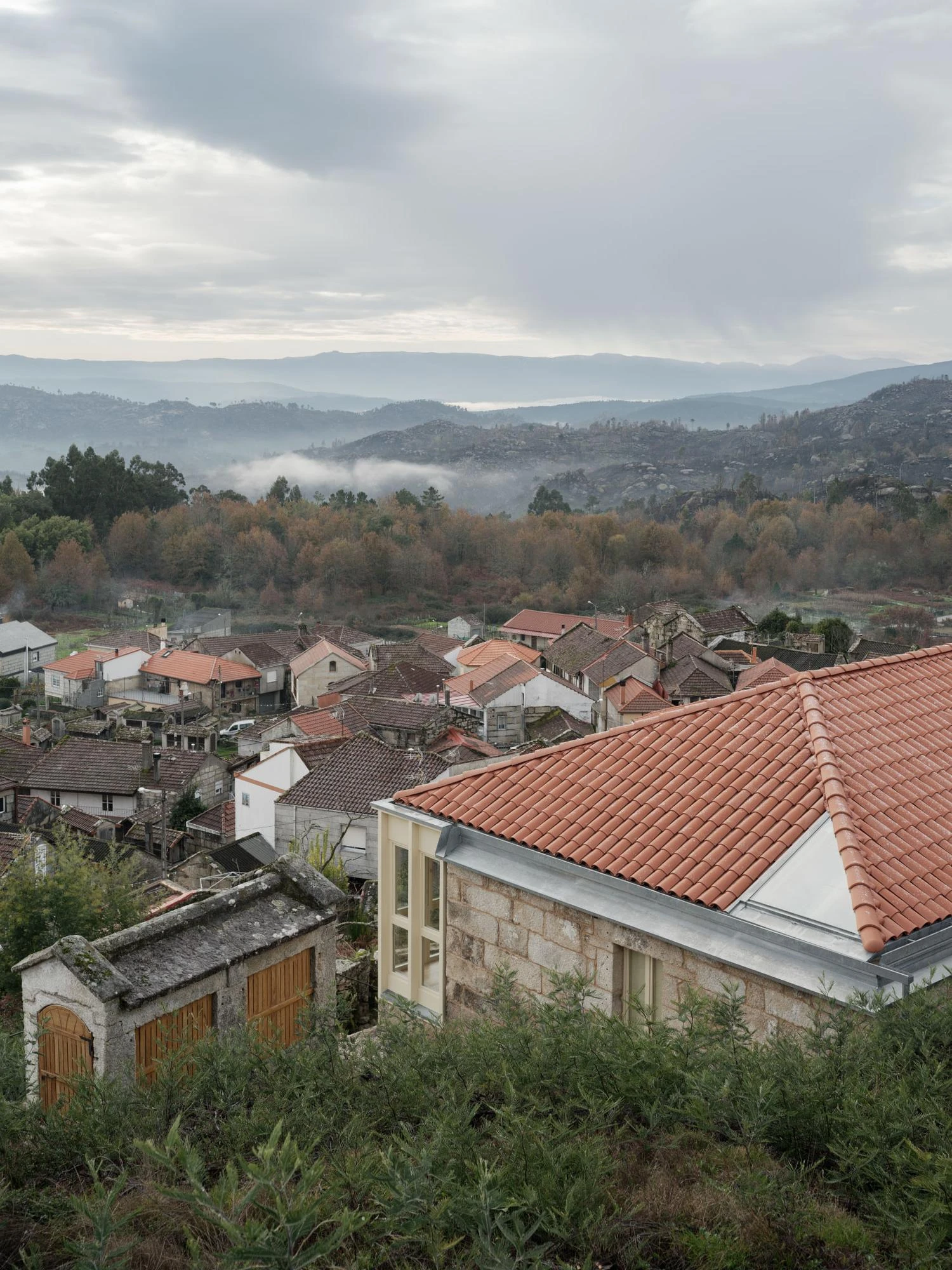Muimenta Multipurpose Social Center by FIRM + BASED, photographed by Luis Díaz Díaz — Seen from above, the new tile roof and horreo settle among the village rooftops under a mist of forested hills.
