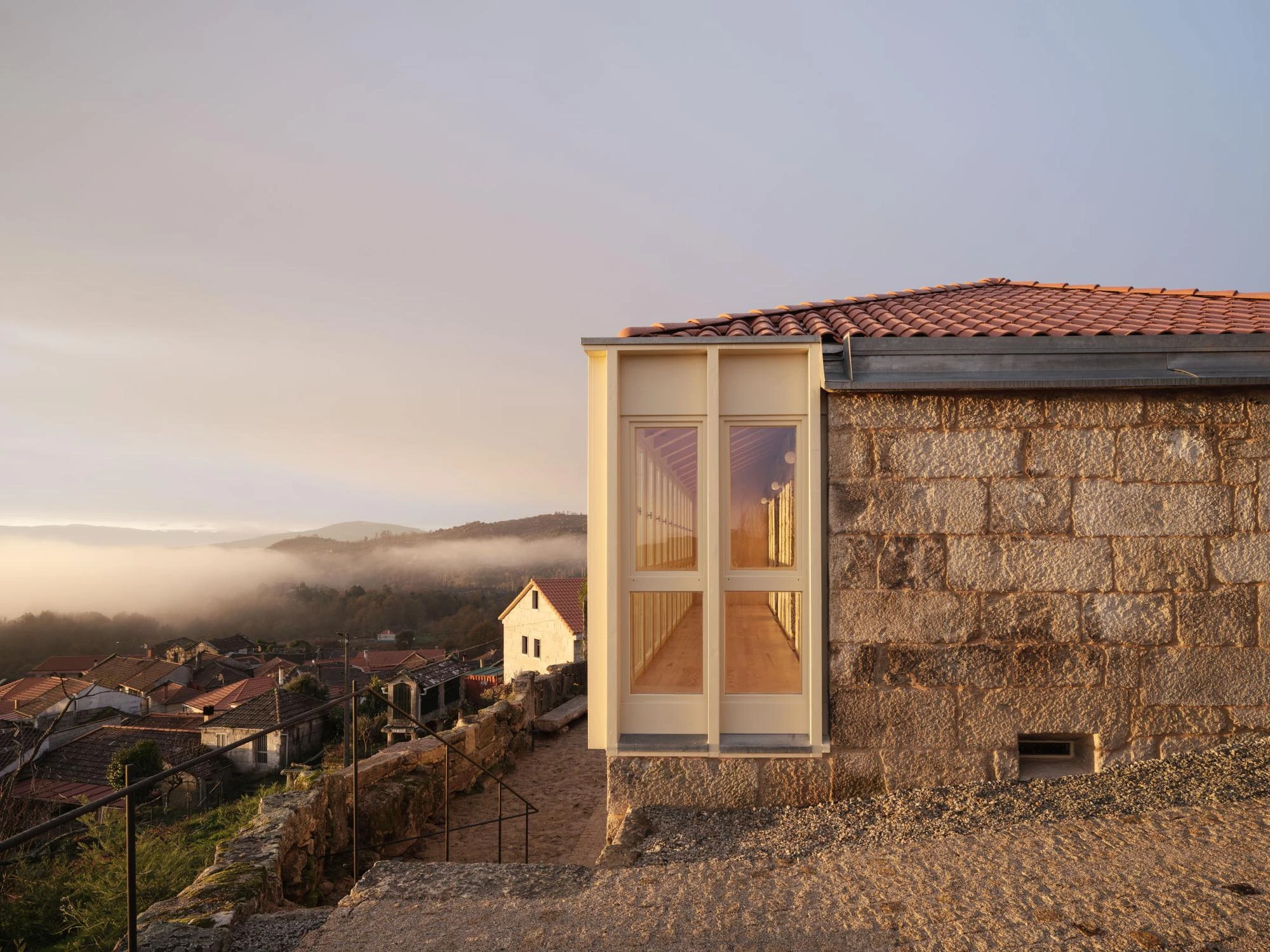 Muimenta Multipurpose Social Center by FIRM + BASED, photographed by Luis Díaz Díaz — At low sun a glazed corner bay catches the light against granite block walls, with the village and valley falling away below.