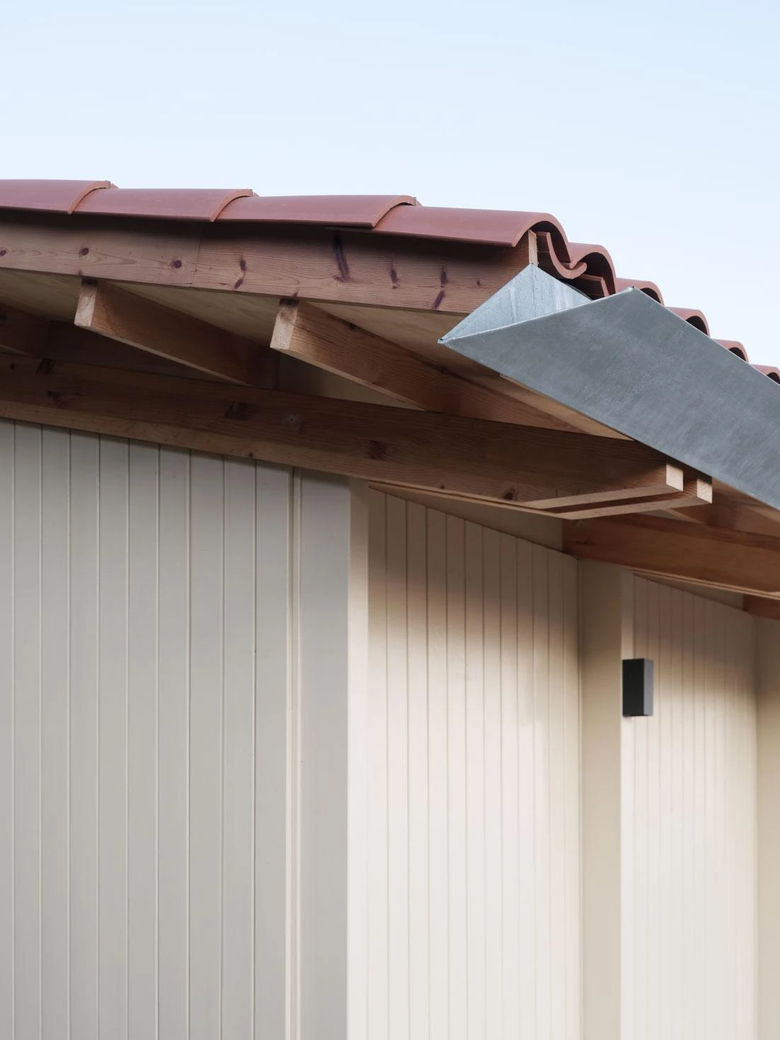 Muimenta Multipurpose Social Center by FIRM + BASED, photographed by Luis Díaz Díaz — A close view of the eave shows tile, exposed timber rafters, a zinc gutter and vertical board cladding.