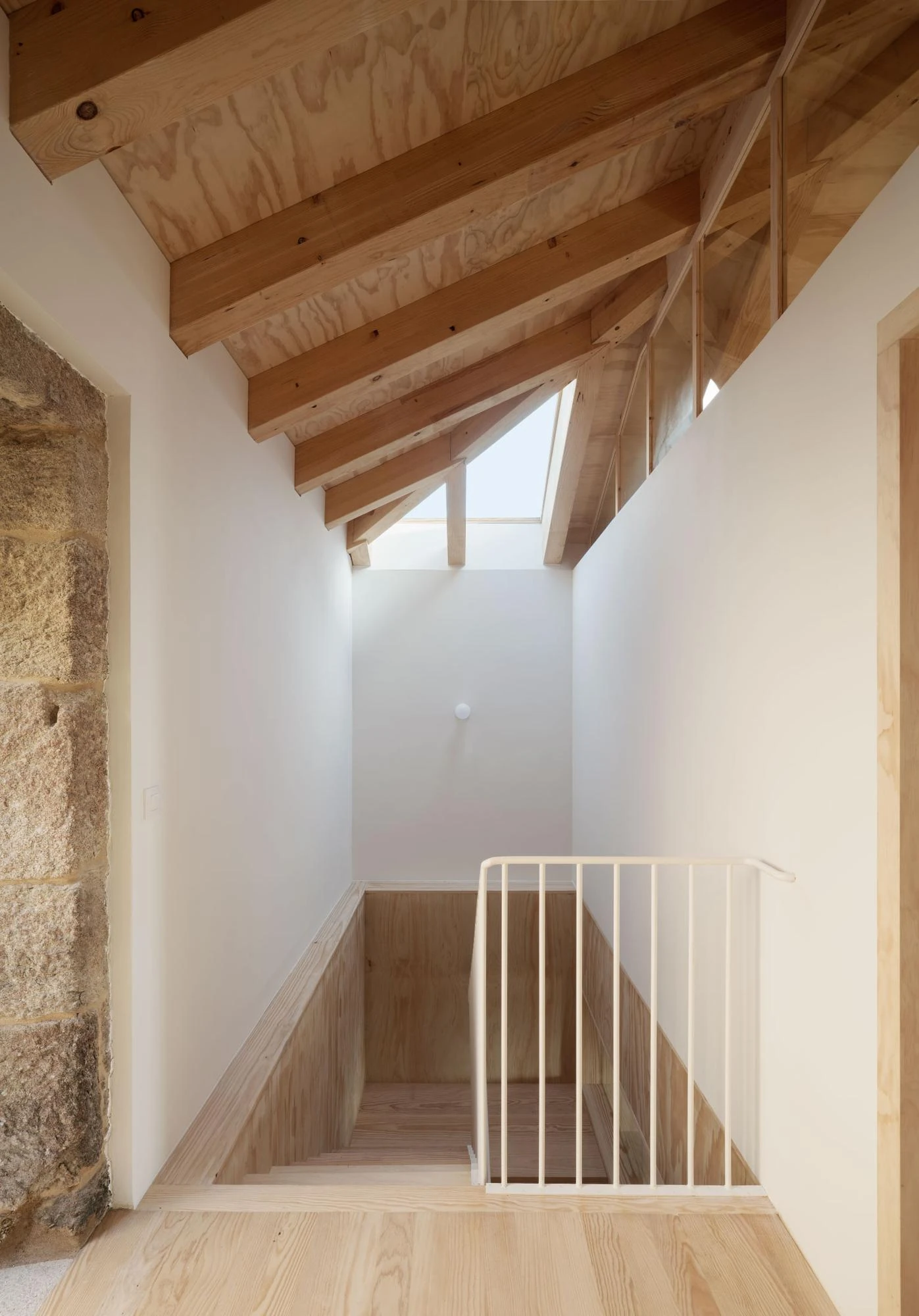 Muimenta Multipurpose Social Center by FIRM + BASED, photographed by Luis Díaz Díaz — Daylight from a roof opening washes down a timber-lined stairwell set against exposed granite masonry.