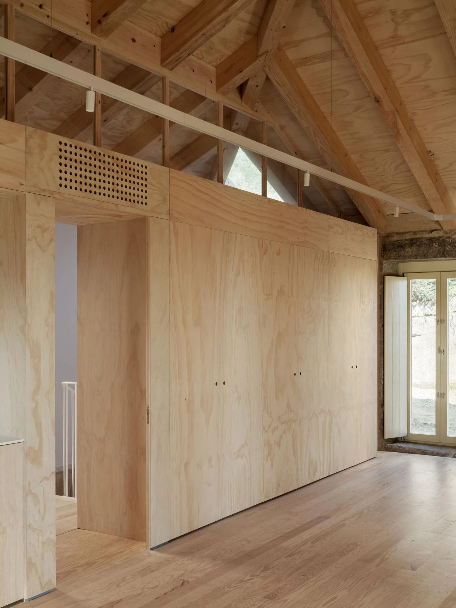 Muimenta Multipurpose Social Center by FIRM + BASED, photographed by Luis Díaz Díaz — A wall of timber joinery lines an open room under an exposed pitched roof, with a glazed door held in the old stone wall.