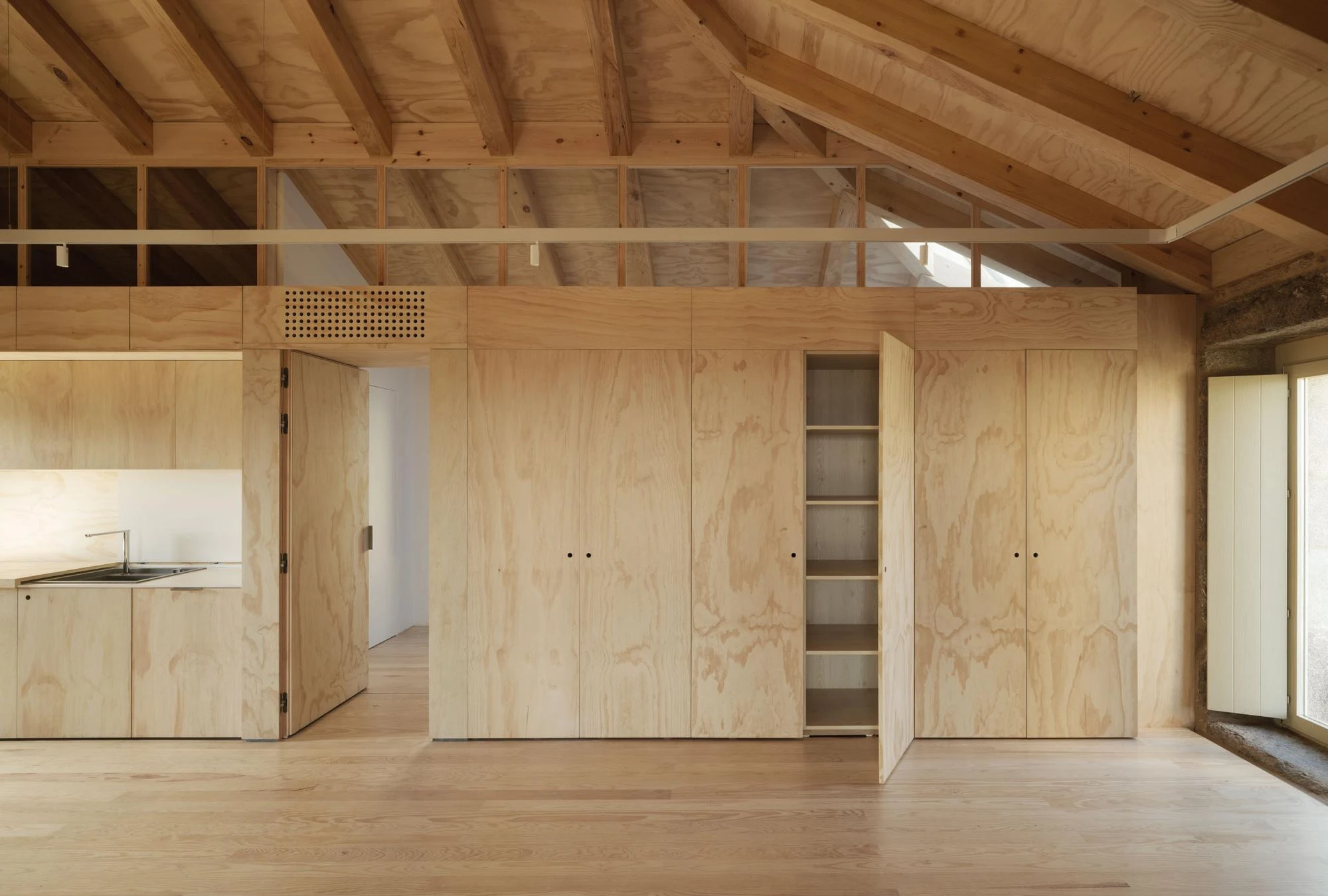 Muimenta Multipurpose Social Center by FIRM + BASED, photographed by Luis Díaz Díaz — A flexible room shows a timber storage wall swung open beside a compact kitchenette and a bay of raw granite.