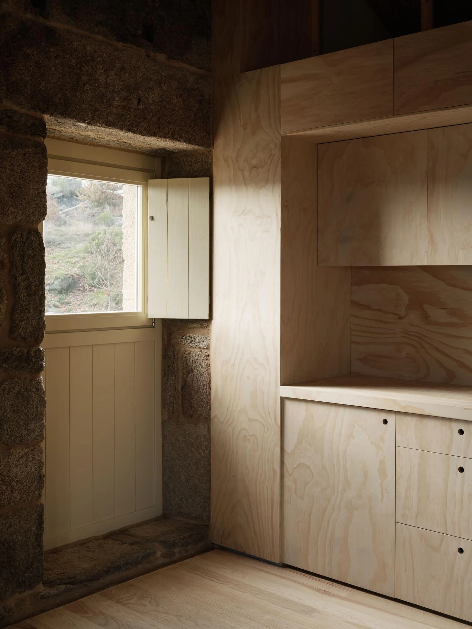 Muimenta Multipurpose Social Center by FIRM + BASED, photographed by Luis Díaz Díaz — A window sits deep in a granite reveal with a timber shutter folded open, next to built-in timber cabinetry.