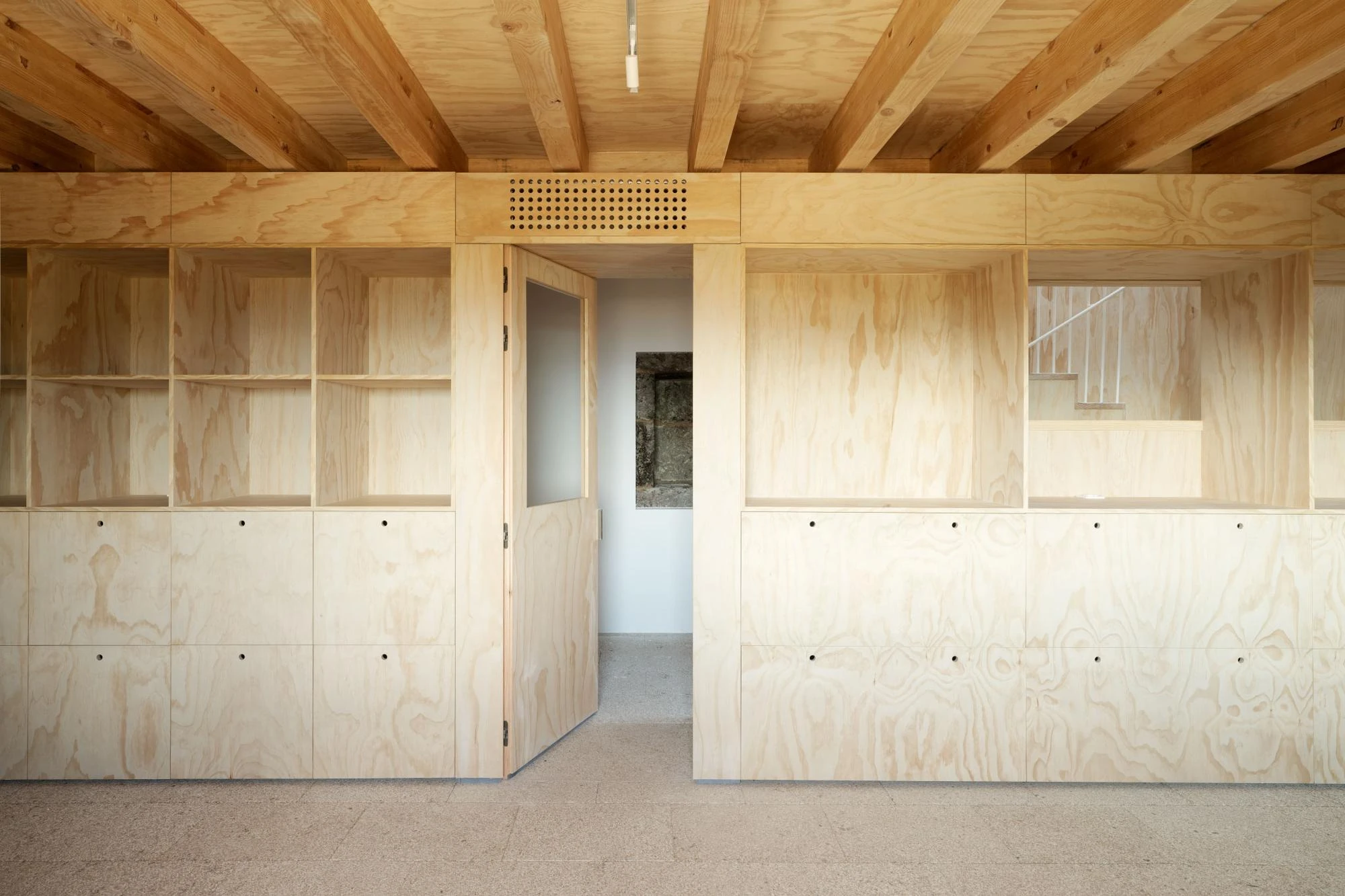 Muimenta Multipurpose Social Center by FIRM + BASED, photographed by Luis Díaz Díaz — Open timber shelving and cabinetry line a room over a granite floor, a doorway giving onto exposed stone.
