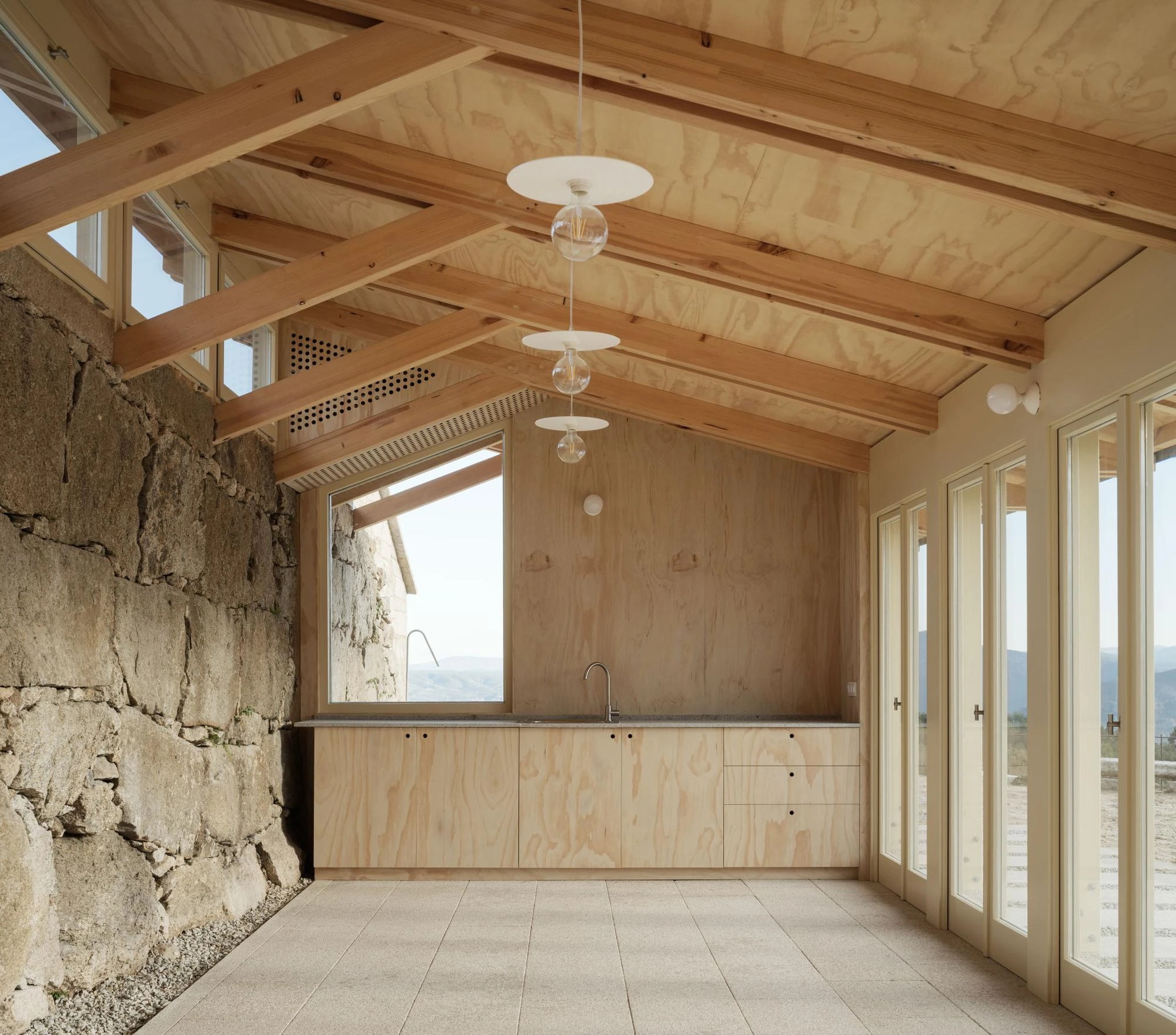 Muimenta Multipurpose Social Center by FIRM + BASED, photographed by Luis Díaz Díaz — Inside the timber pavilion, an exposed rafter roof runs above a counter set against the old granite wall, glazing opening to the valley.