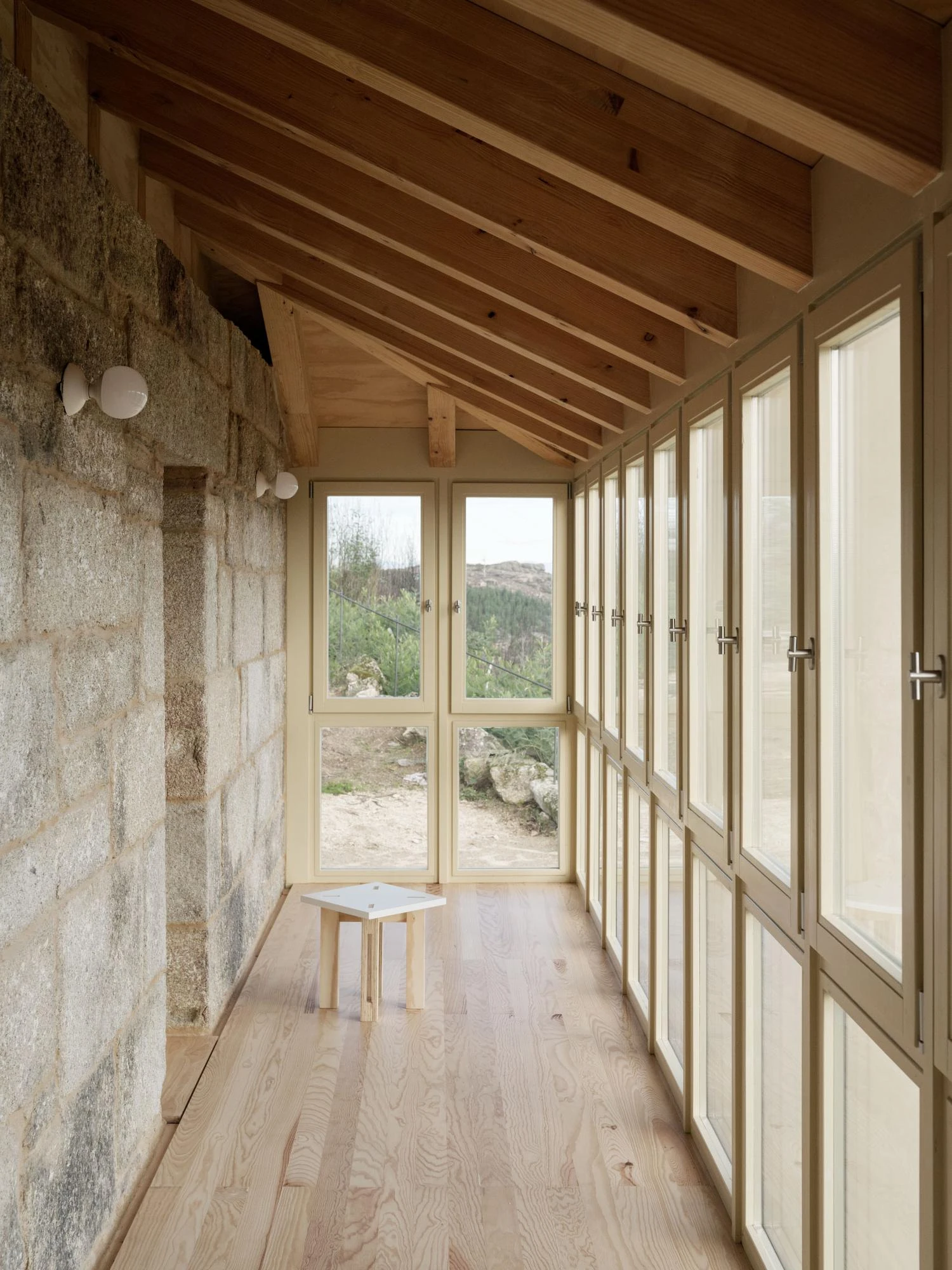 Muimenta Multipurpose Social Center by FIRM + BASED, photographed by Luis Díaz Díaz — A glazed gallery runs along a granite wall over a timber floor, a small stool set before the mullioned windows.