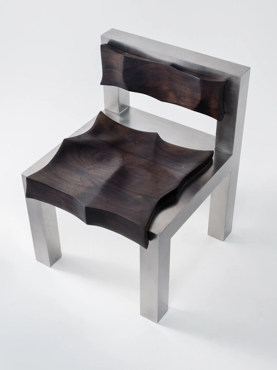 Soft Tensions by Studio Sija, photographed by Pēteris Vīksna — A three-quarter view of the full chair, its dark walnut seat and low back scooped into shallow basins between square-section legs of brushed stainless steel.