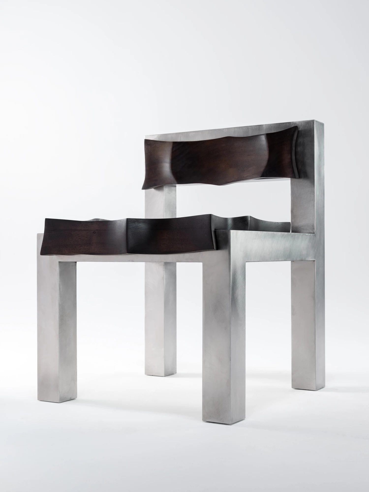 Soft Tensions by Studio Sija, photographed by Pēteris Vīksna — A three-quarter rear view with the walnut seat and backrest floating as two dark carved panels inside the welded steel frame.