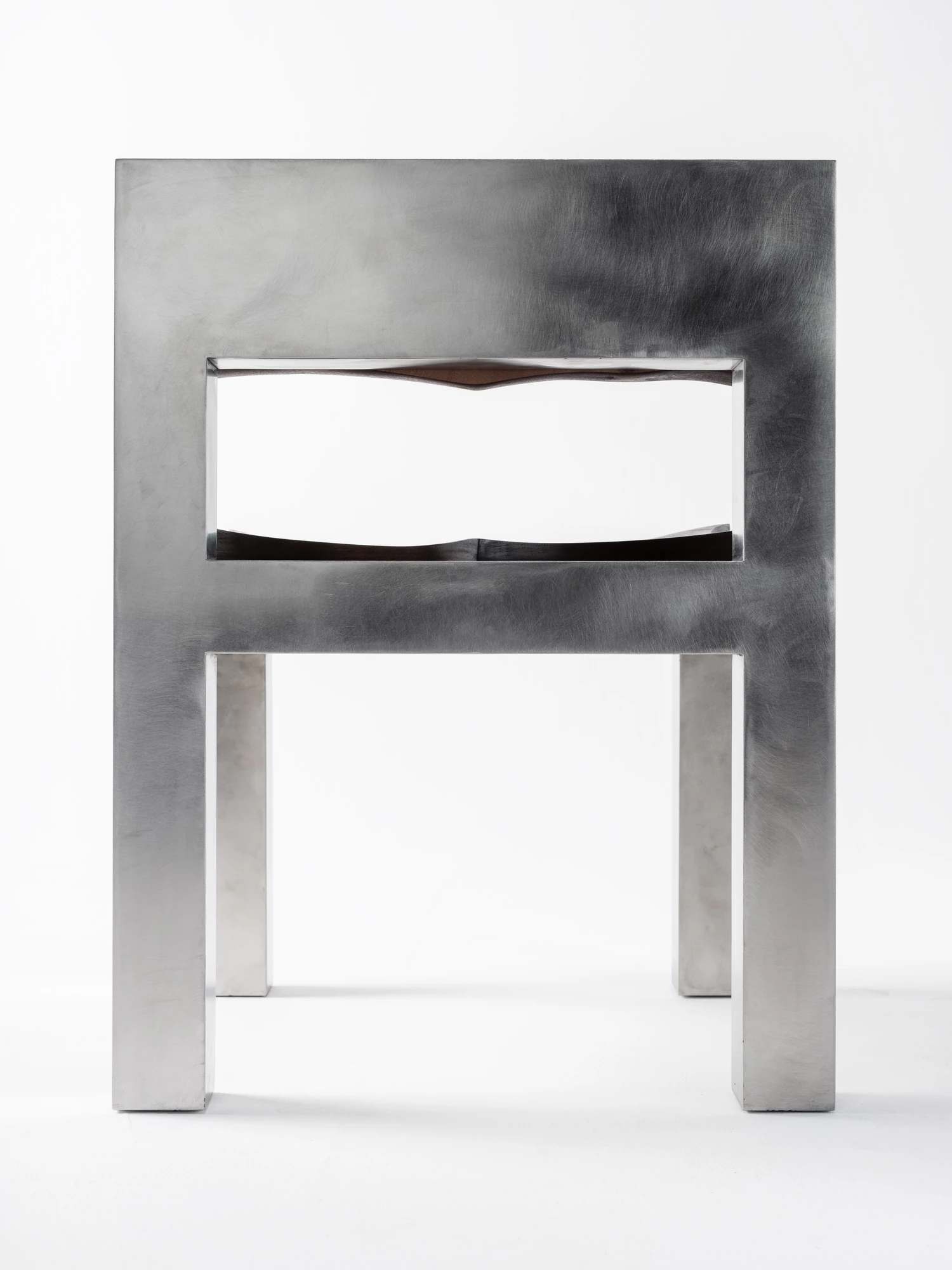 Soft Tensions by Studio Sija, photographed by Pēteris Vīksna — A frontal view through the open steel frame, the carved wooden seat and back reading as two dark bands set within the metal rectangle.