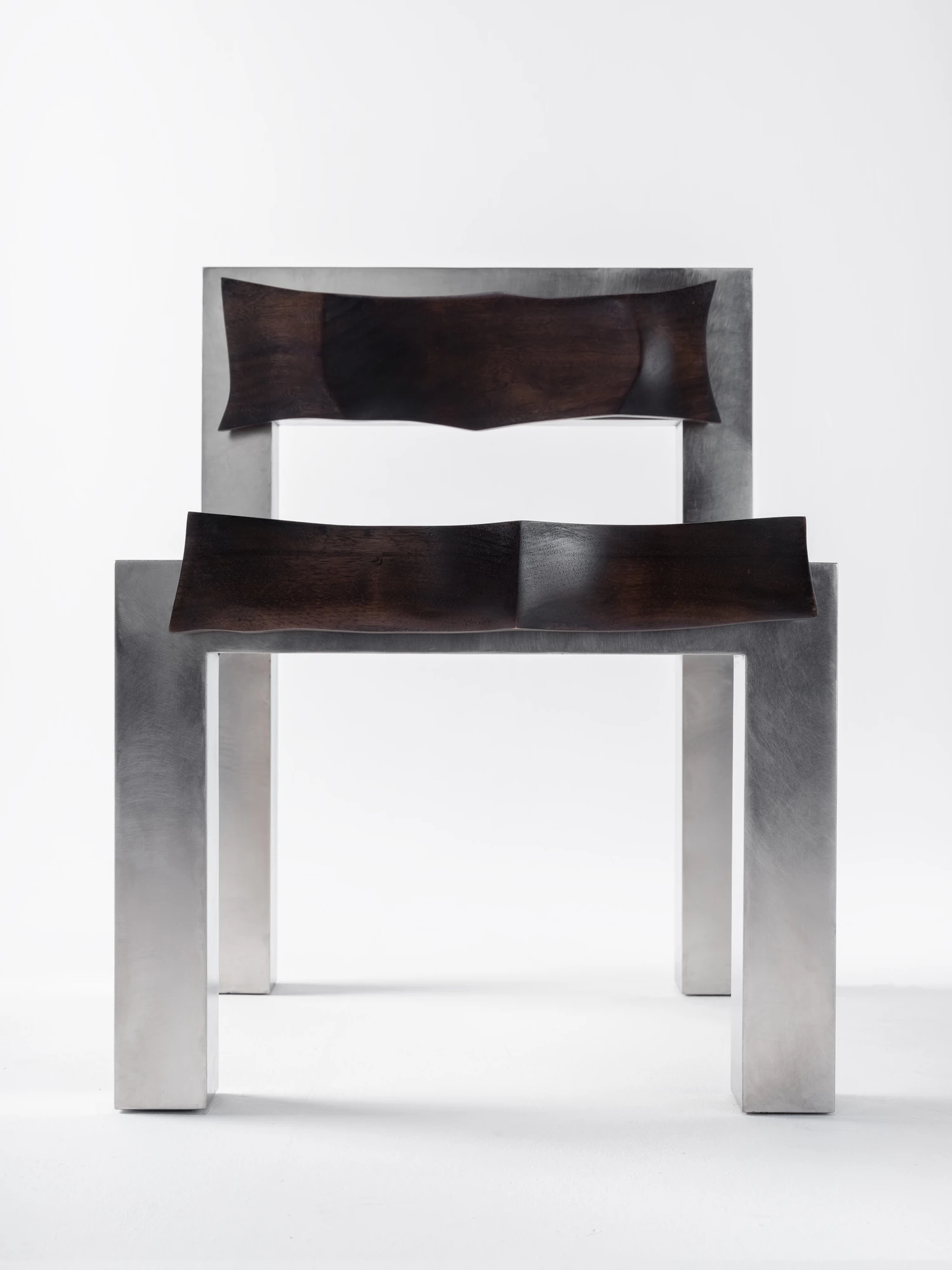 Soft Tensions by Studio Sija, photographed by Pēteris Vīksna — The chair seen straight on, back and seat panels of dark walnut carved to pinched waisted edges within the stainless steel structure.