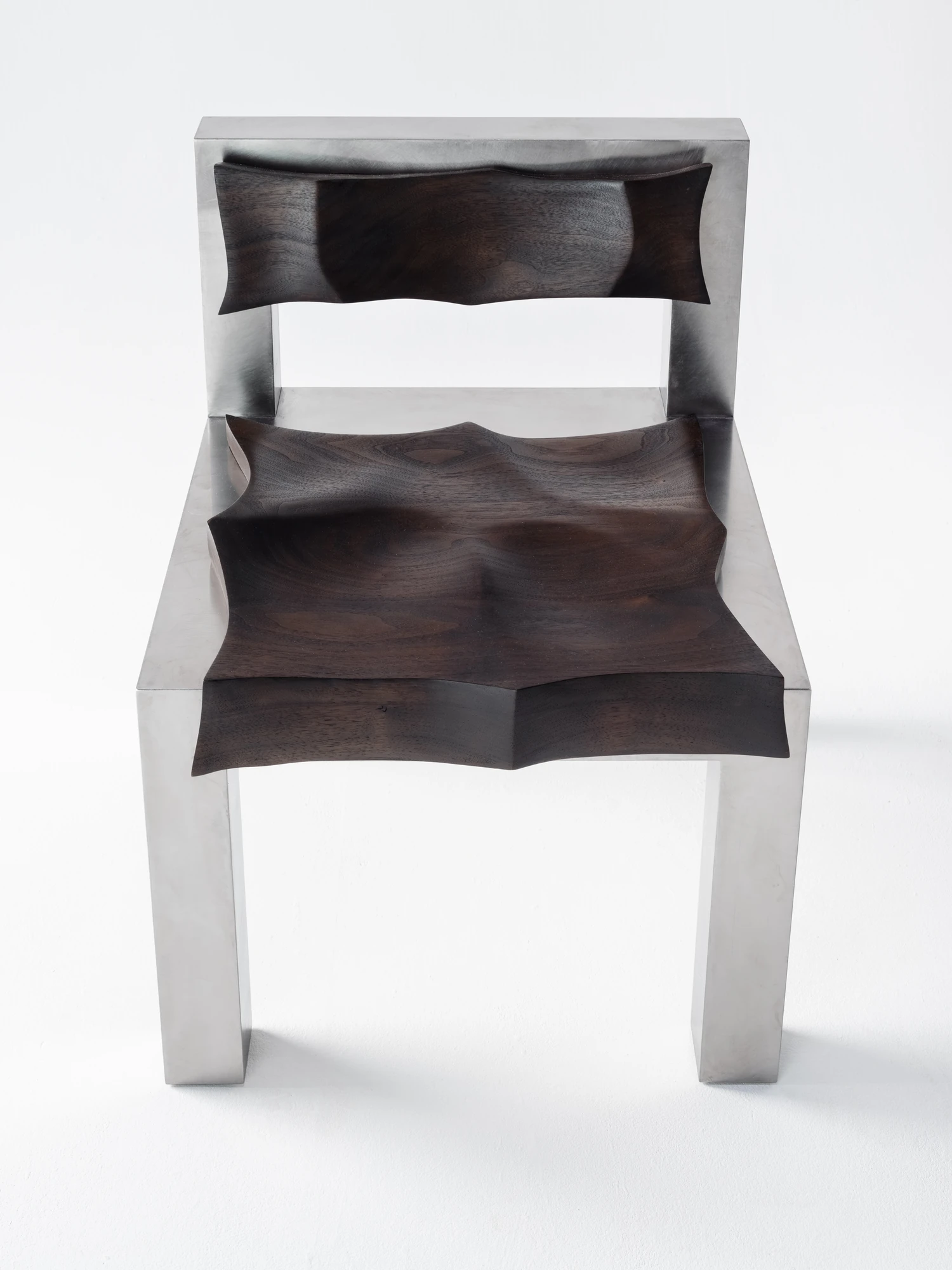 Soft Tensions by Studio Sija, photographed by Pēteris Vīksna — A low front angle showing the deeply scooped walnut seat and matching backrest set into the brushed metal frame.