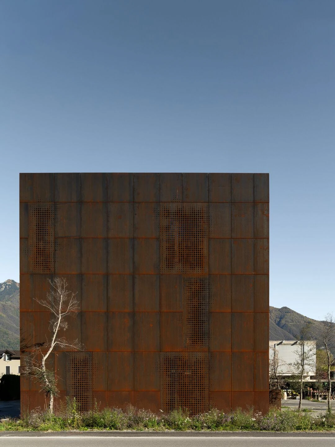 Urs Hauser Winery by WDMRA, photographed by Giacomo Albo — A tall frontal view from the road, the weathered steel face rising over a wildflower verge with mountains behind.