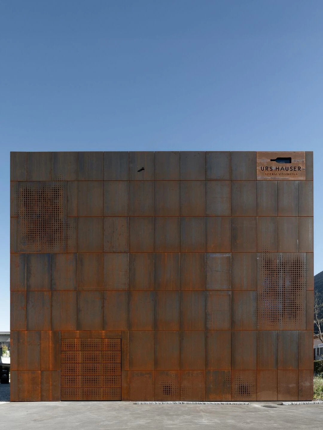 Urs Hauser Winery by WDMRA, photographed by Giacomo Albo — The facade carrying a cut-steel bottle sign and the name Urs Hauser, scattered perforated panels reading against open sky.