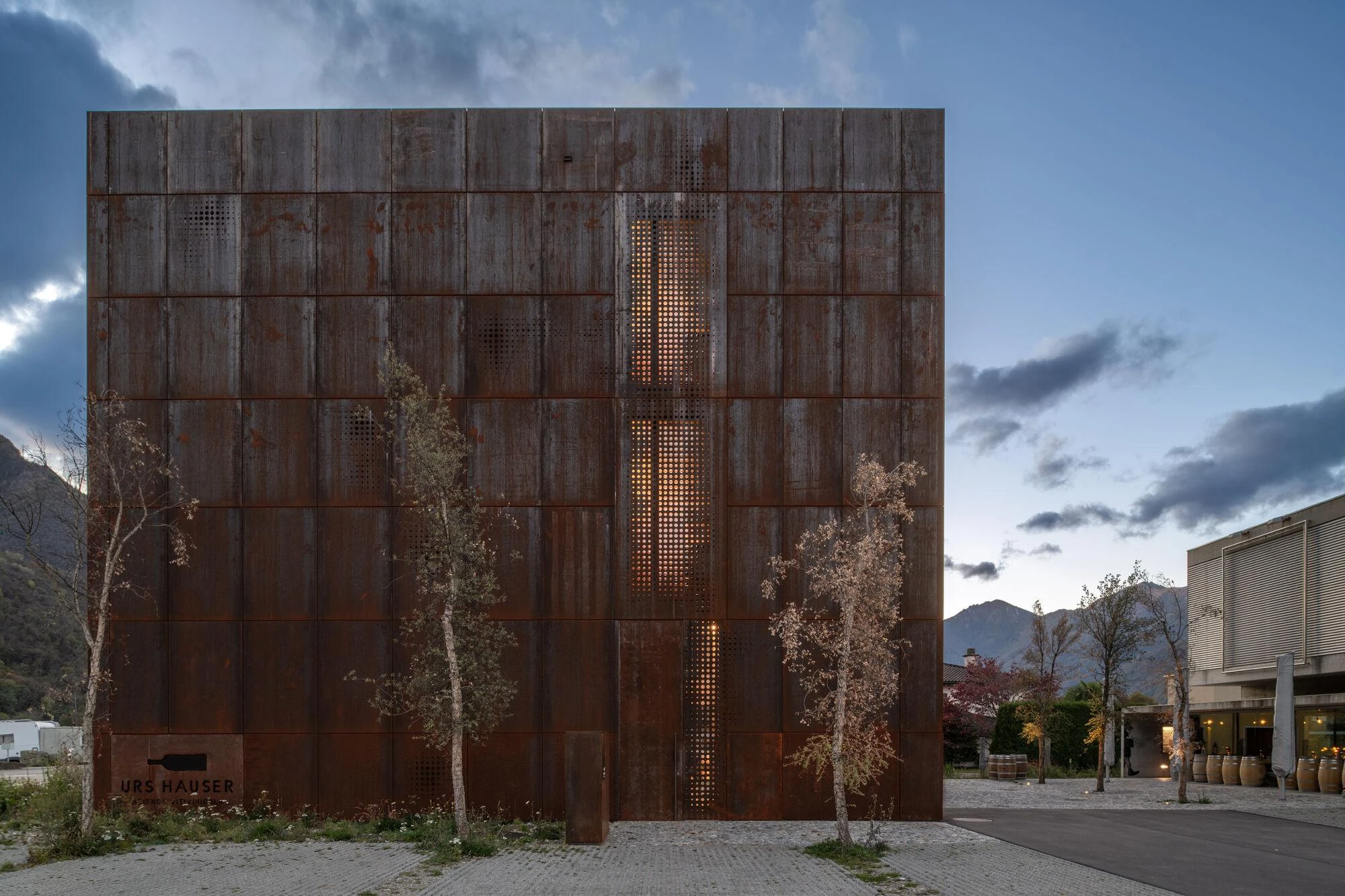Urs Hauser Winery by WDMRA, photographed by Giacomo Albo — At dusk a perforated column glows warm from within the steel, the lit neighbouring building visible to the right.