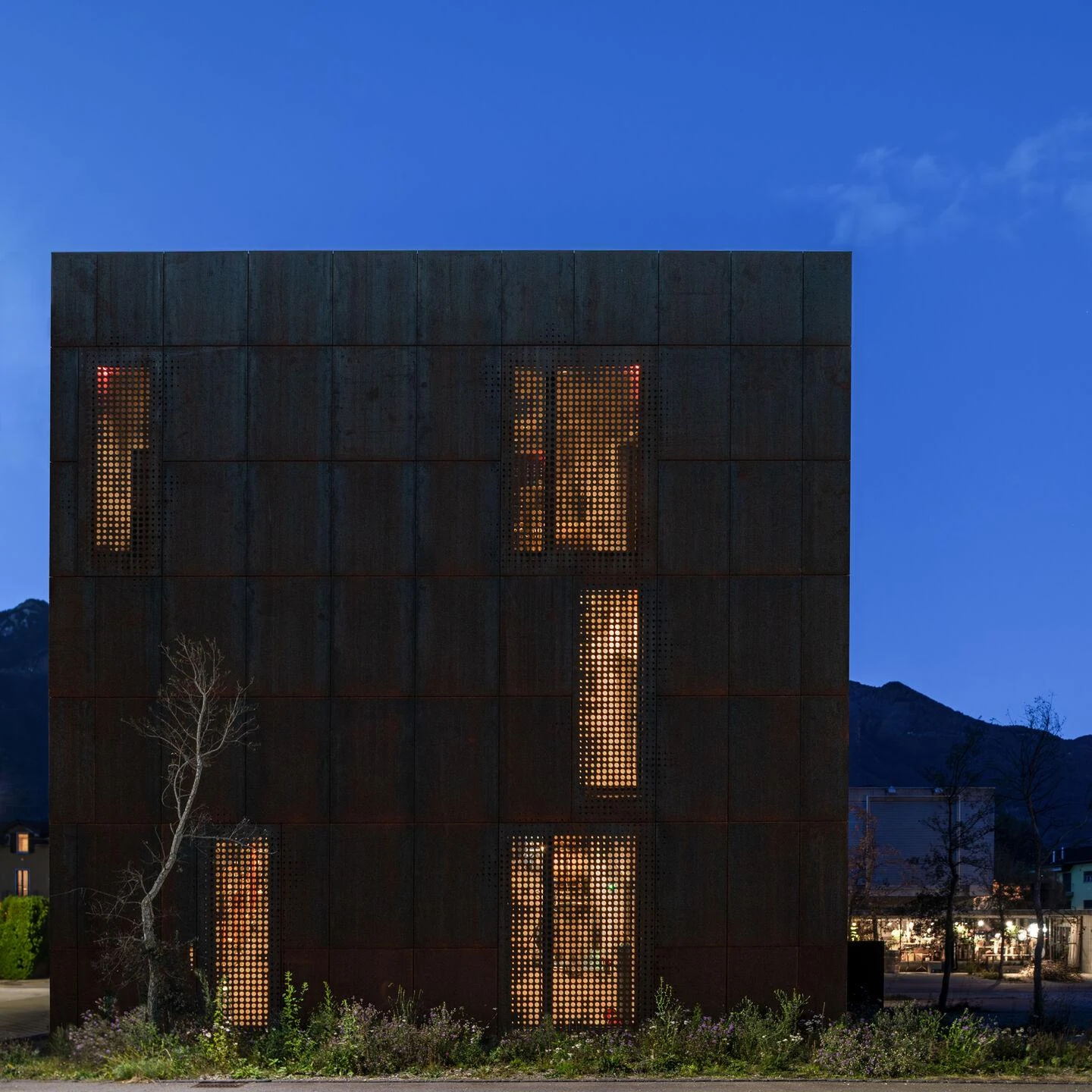 Urs Hauser Winery by WDMRA, photographed by Giacomo Albo — Blue-hour elevation with amber light pooling behind perforated panels like windows scattered across the dark facade.