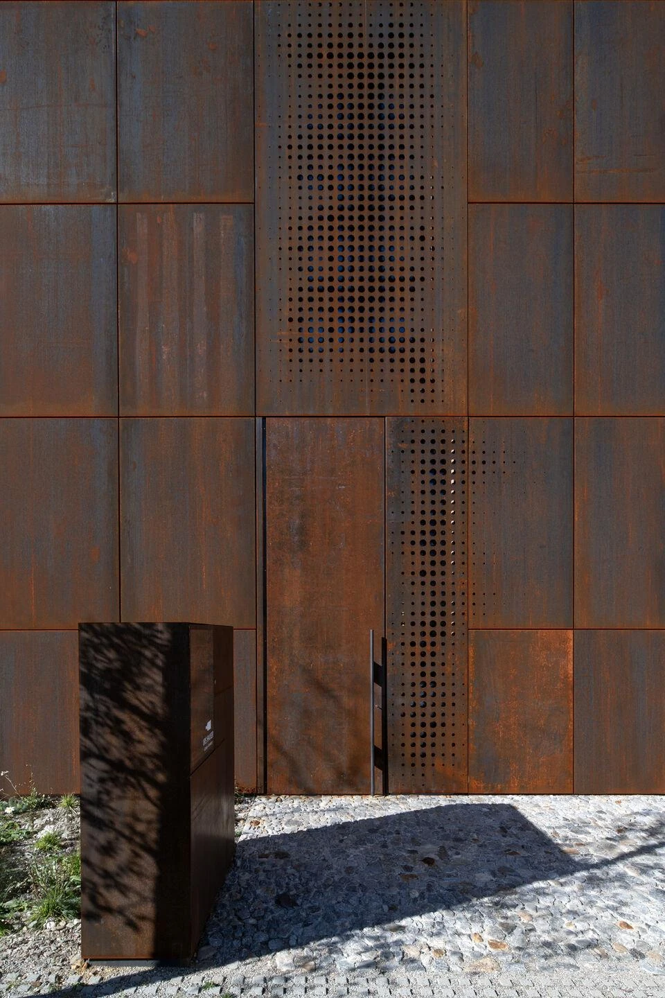 Urs Hauser Winery by WDMRA, photographed by Giacomo Albo — The base of the facade with a freestanding steel box beside a perforated door band, cobblestones underfoot.