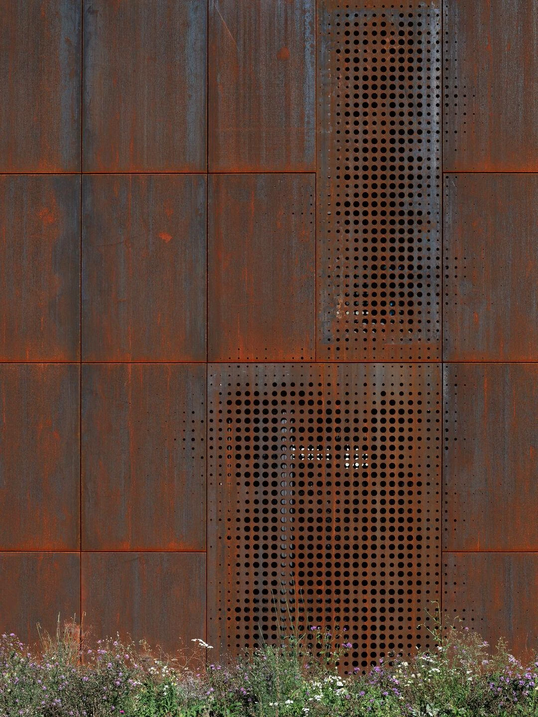 Urs Hauser Winery by WDMRA, photographed by Giacomo Albo — A close view of the perforated Corten, holes fading from dense to sparse, wildflowers gathered at the foot of the wall.