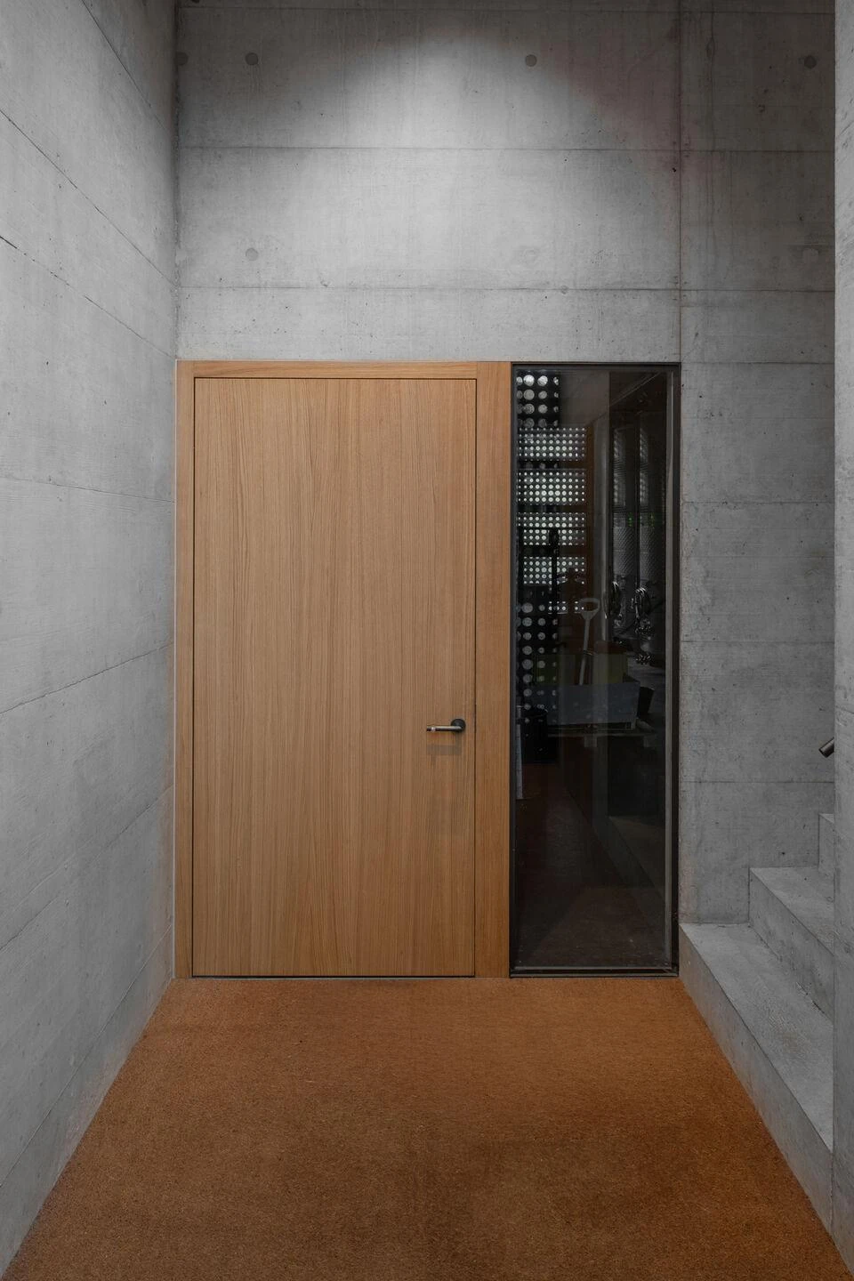 Urs Hauser Winery by WDMRA, photographed by Giacomo Albo — An interior concrete lobby, an oak door set beside a glazed slot onto the tank hall, a cork-coloured floor below.