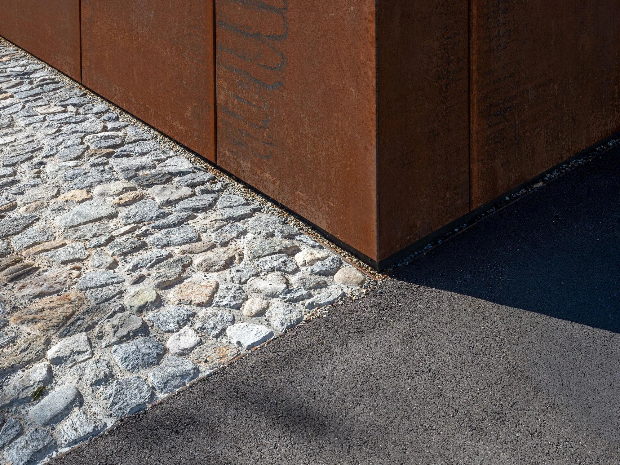 Urs Hauser Winery by WDMRA, photographed by Giacomo Albo — The base corner where cobblestone paving, gravel and asphalt meet the steel wall.