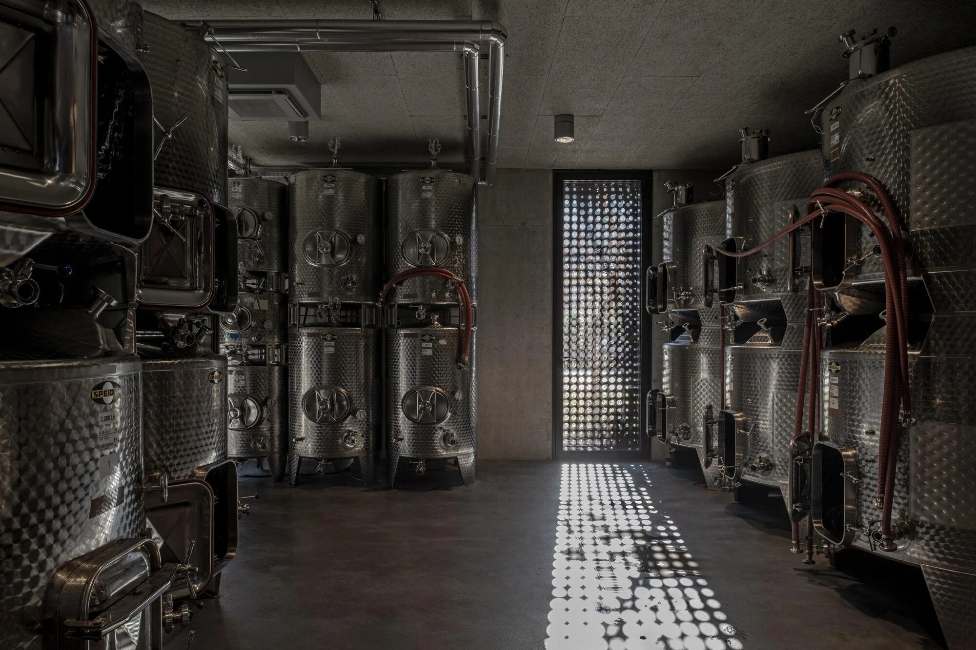 Urs Hauser Winery by WDMRA, photographed by Giacomo Albo — Stainless fermentation tanks in a concrete hall, a perforated door throwing a column of dotted light across the floor.