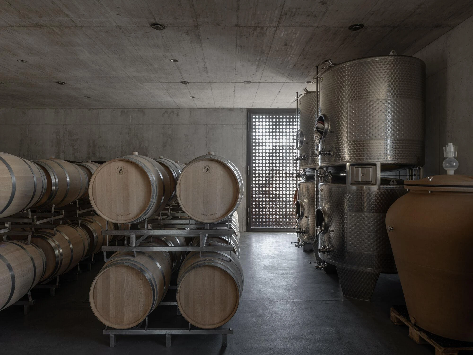 Urs Hauser Winery by WDMRA, photographed by Giacomo Albo — The barrel cellar with oak barrels and steel tanks alongside a terracotta amphora, the perforated door glowing behind them.