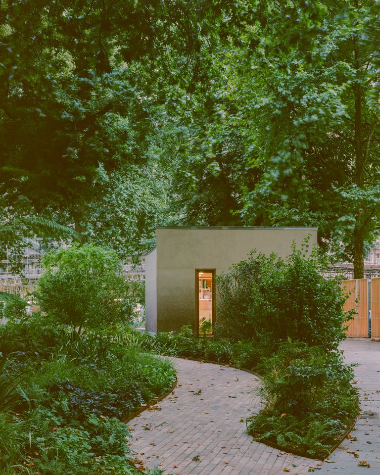 Finsbury Circus Parks Office by Studio Weave, photographed by Lorenzo Zandri — A curved corner of the clay-render volume meets a brick paver path winding through ferns and shrubs, a doorway glowing warm within.