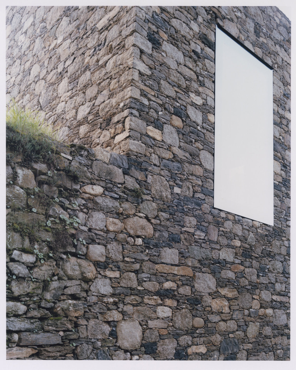 Office Orma Architettura by Orma Architettura — A close view of the stone corner, a trapezoidal window set flush in the wall above a mossed ruined base.