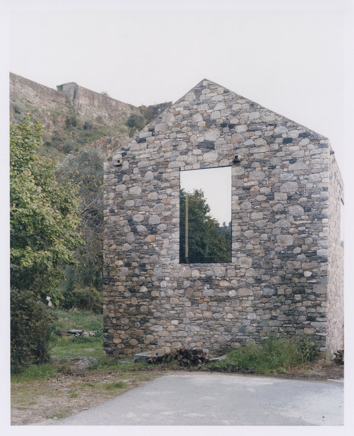Office Orma Architettura by Orma Architettura — The gabled stone volume seen head-on, a single tall window set into the gable wall, the ruined citadel rampart on the ridge above.