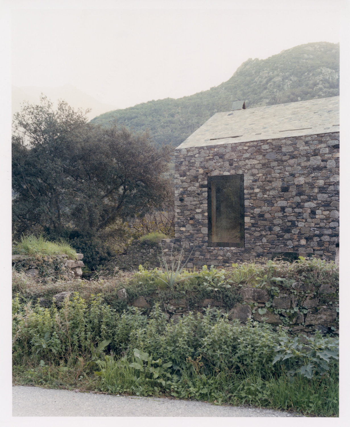 Office Orma Architettura by Orma Architettura — The long stone flank with a single dark window, low ruined garden walls in front and misted mountains behind.