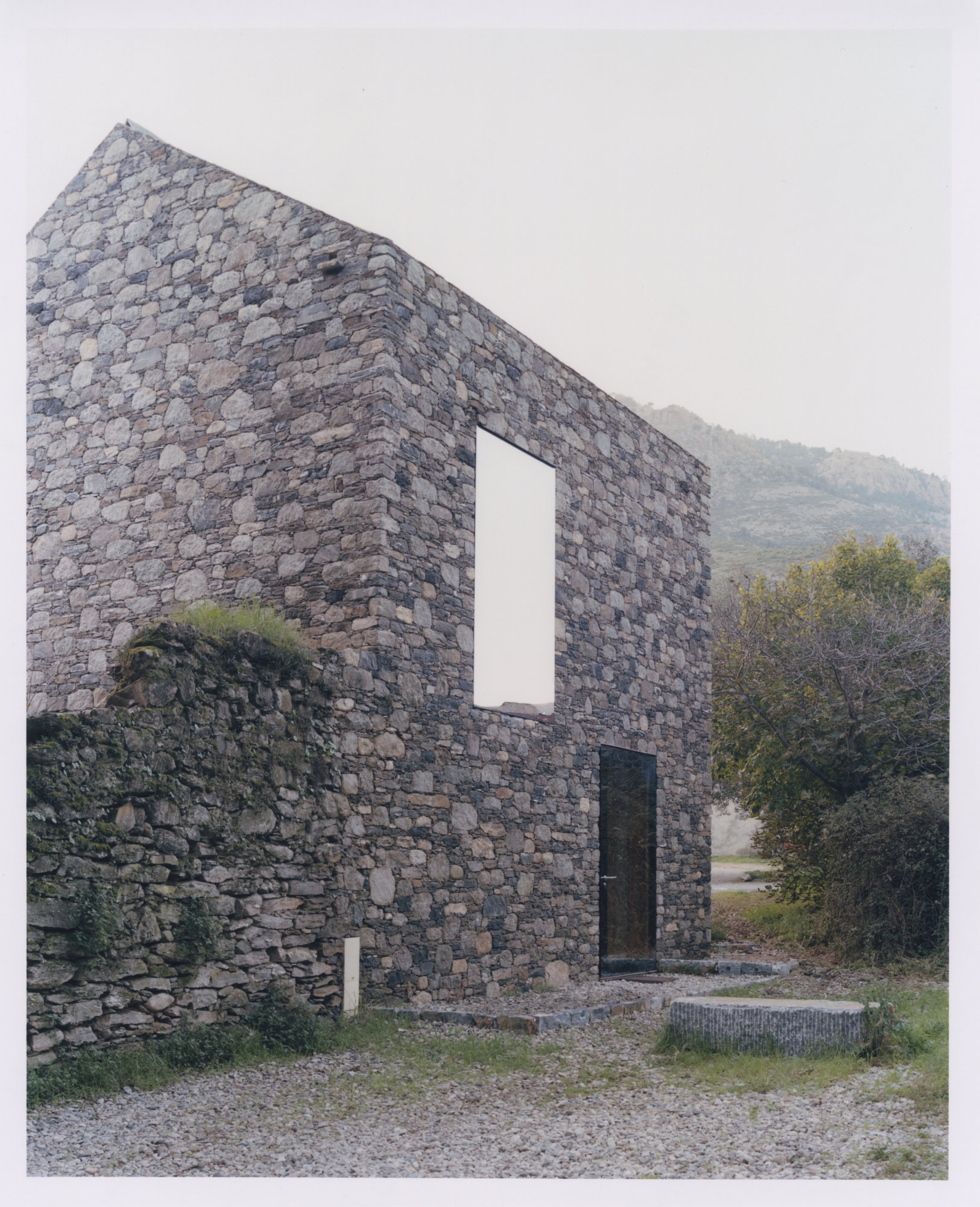 Office Orma Architettura by Orma Architettura — The tall gable end with a vertical window and a dark doorway, a mossed ruined wall to the left and mountains beyond.