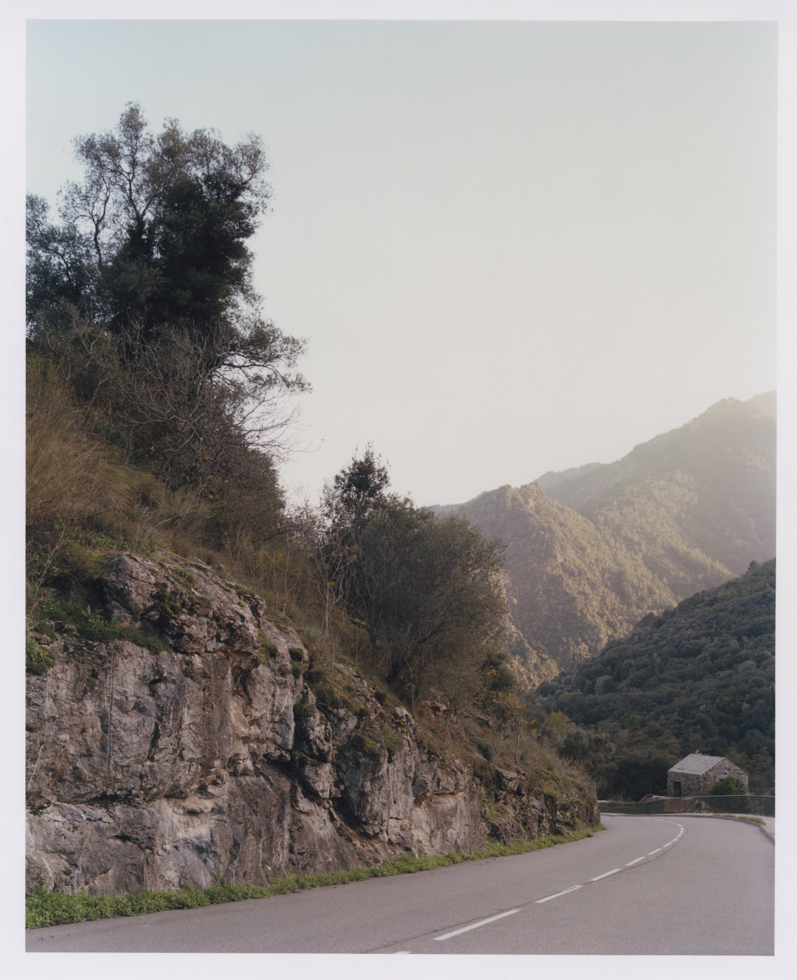 Office Orma Architettura by Orma Architettura — A mountain road curves past a rock outcrop, the small stone building visible at the far right below wooded slopes.