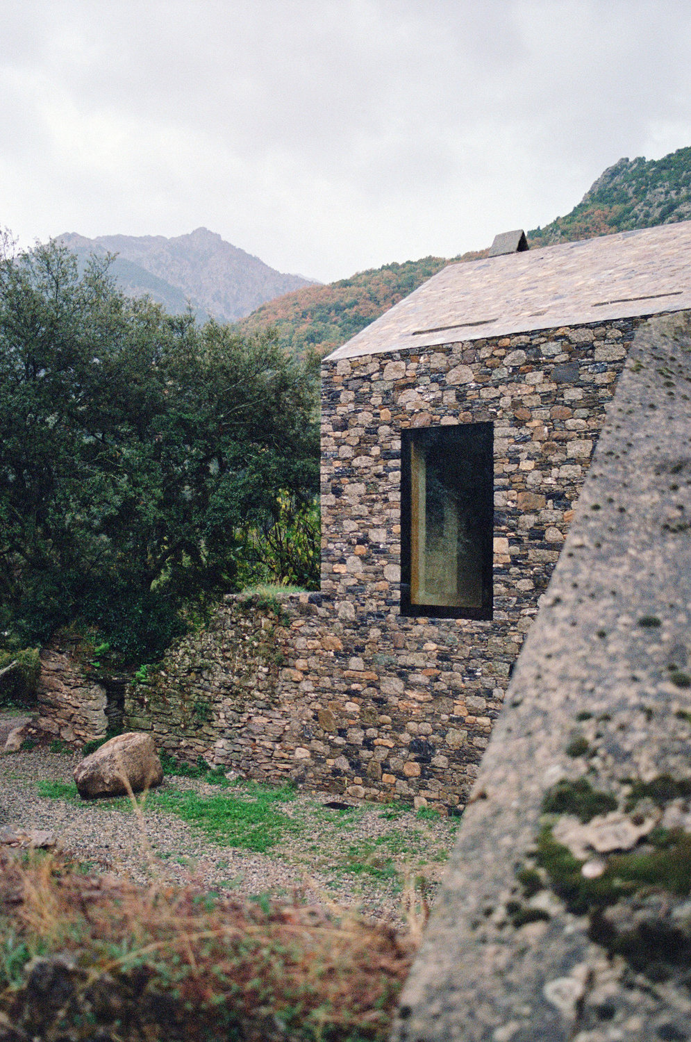 Office Orma Architettura by Orma Architettura — The stone volume from the entrance side, its raked roofline and a tall window above a gravel forecourt.