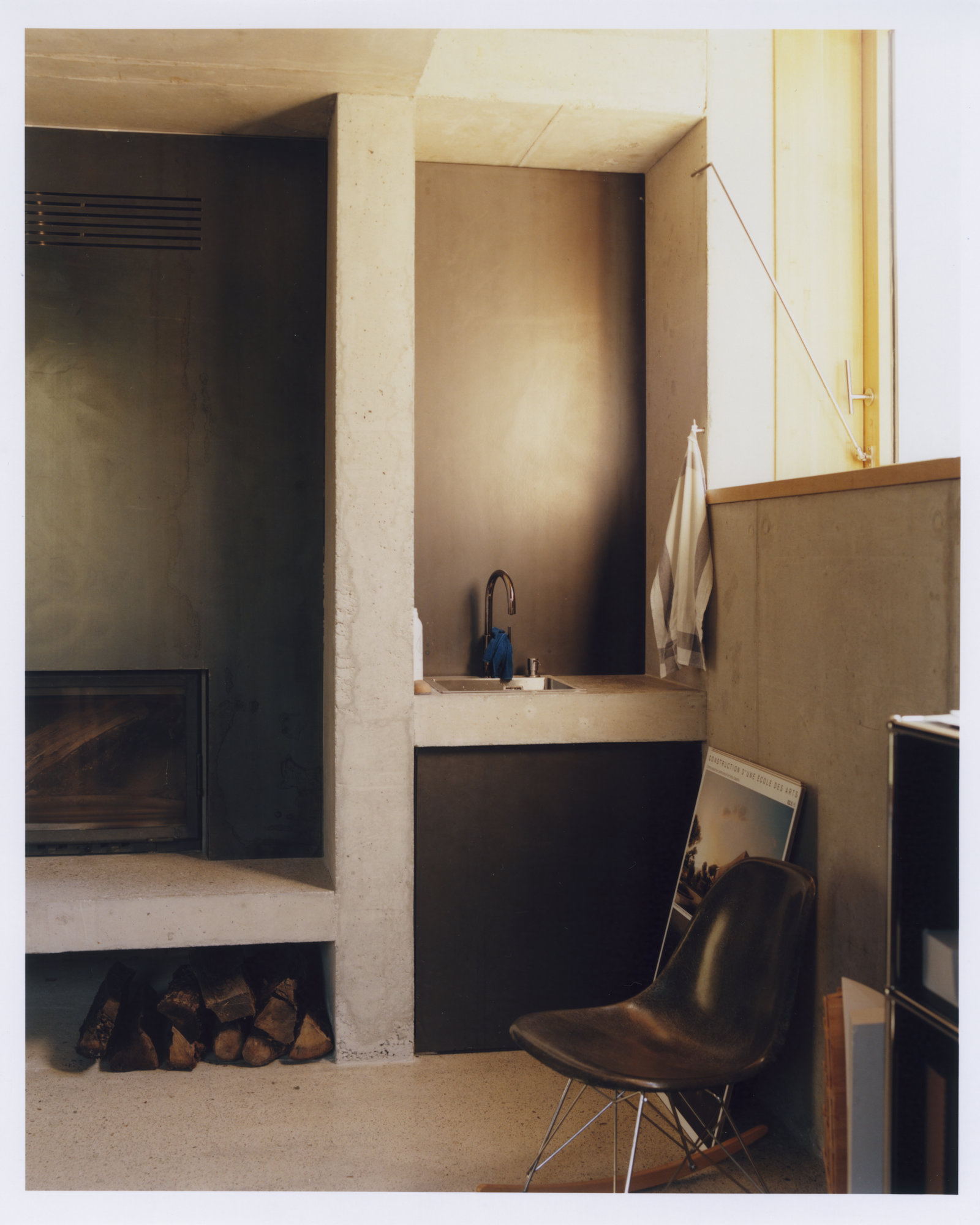Office Orma Architettura by Orma Architettura — An interior corner in board-formed concrete, a cast concrete sink niche, a recessed hearth stacked with logs and a black chair.