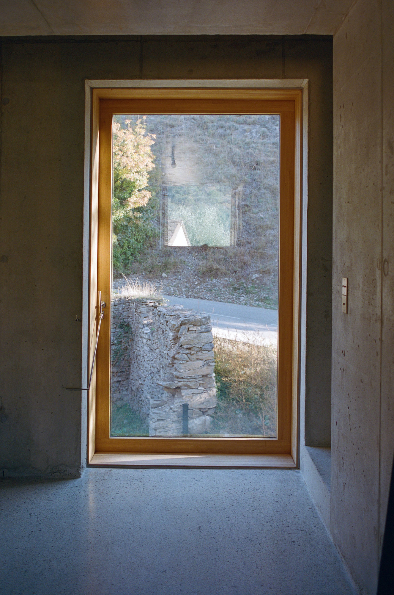Office Orma Architettura by Orma Architettura — A deep timber-framed window in a concrete wall, looking out to a ruined stone wall and the road below.
