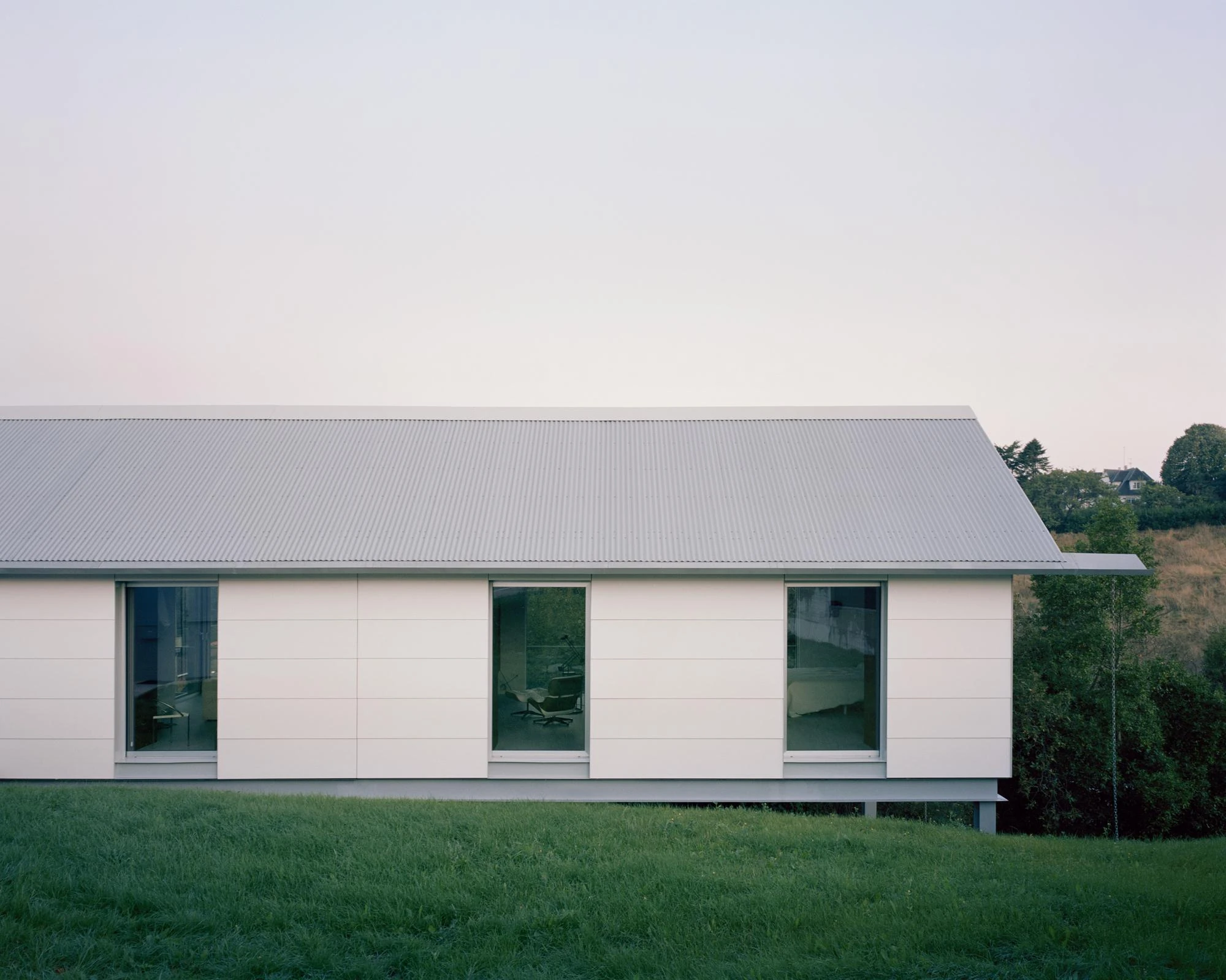 Casa Guadalupe by HANGHAR, photographed by Rory Gardiner — The long white flank of Casa Guadalupe, clad in fiber-cement sandwich panels under a corrugated metal roof, stands a step above a cut lawn at dusk.