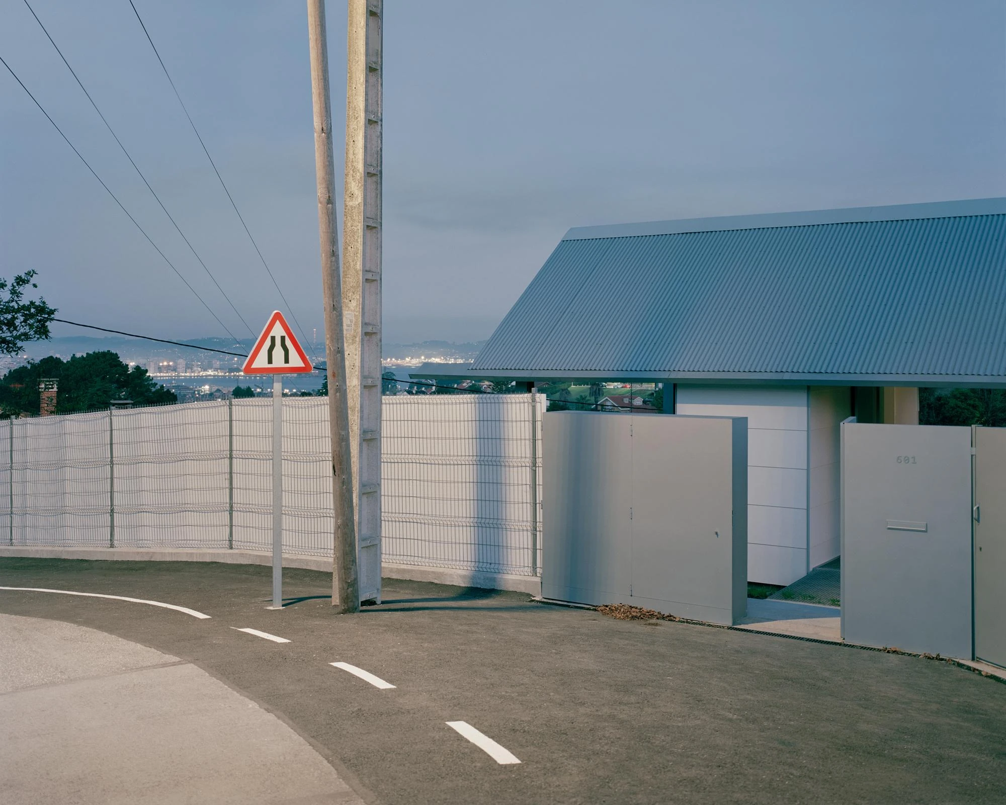 Casa Guadalupe by HANGHAR, photographed by Rory Gardiner — Dusk at the street approach, where a curving lane and a wire fence meet the house's grey sliding panels and corrugated roof.