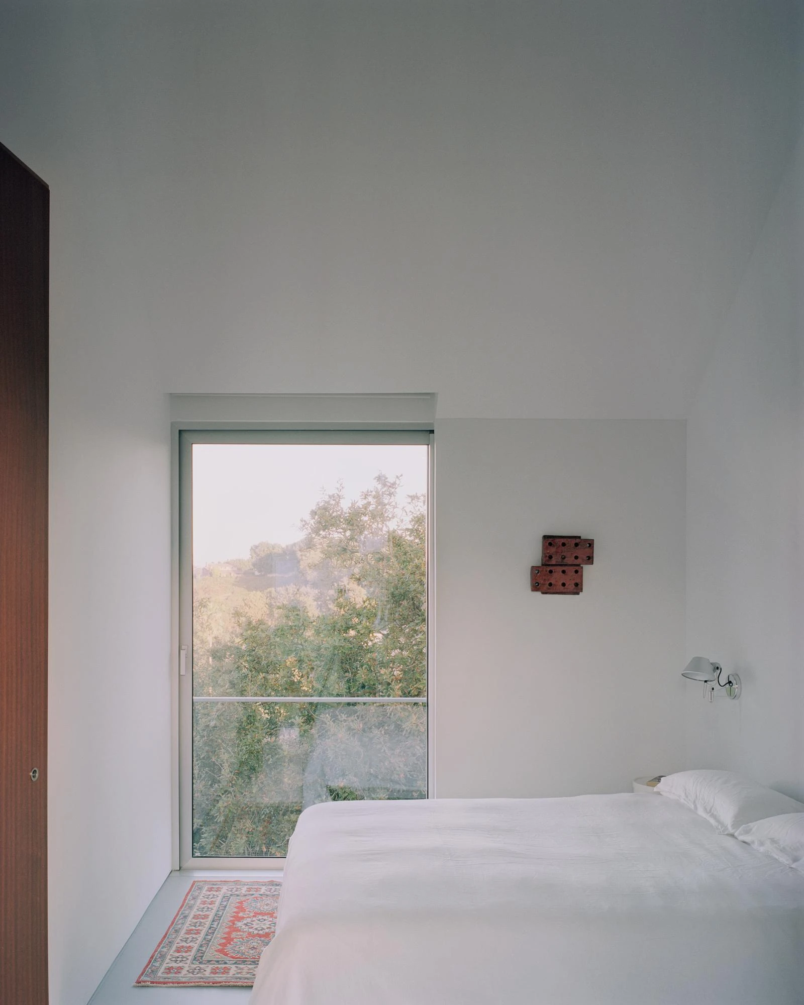 Casa Guadalupe by HANGHAR, photographed by Rory Gardiner — A bedroom with white walls and a tall window framing wooded hillside.