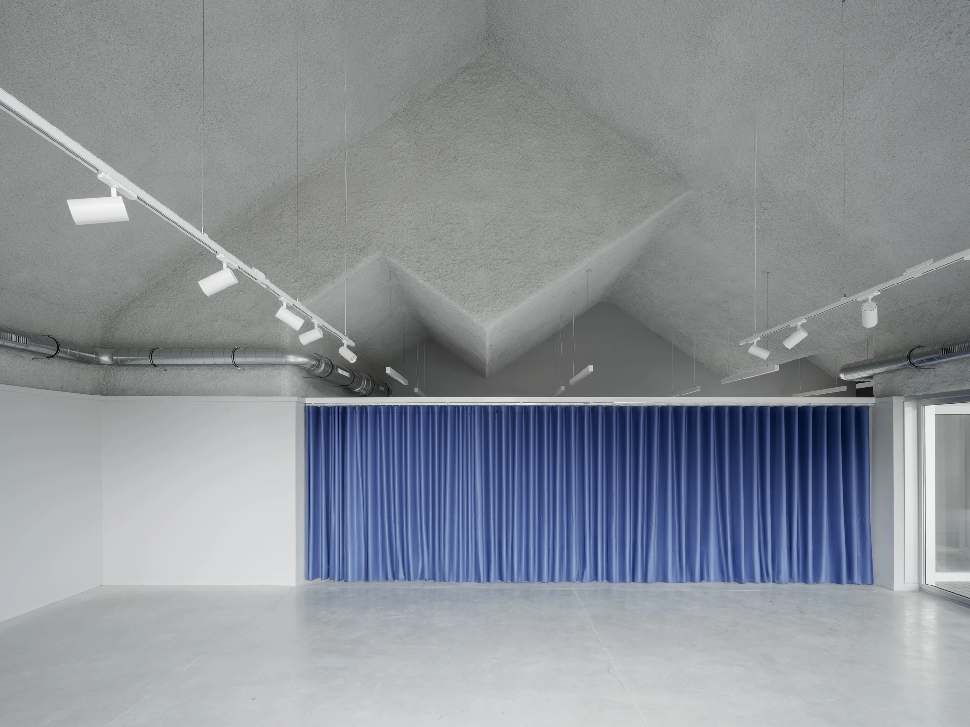 Goians Community Center by NULA.STUDIO + FCA, photographed by Luis Díaz Díaz / NULA.STUDIO — A full wall of blue velvet curtain hangs beneath the folded grey rockwool ceiling, the polished concrete floor reflecting the light.