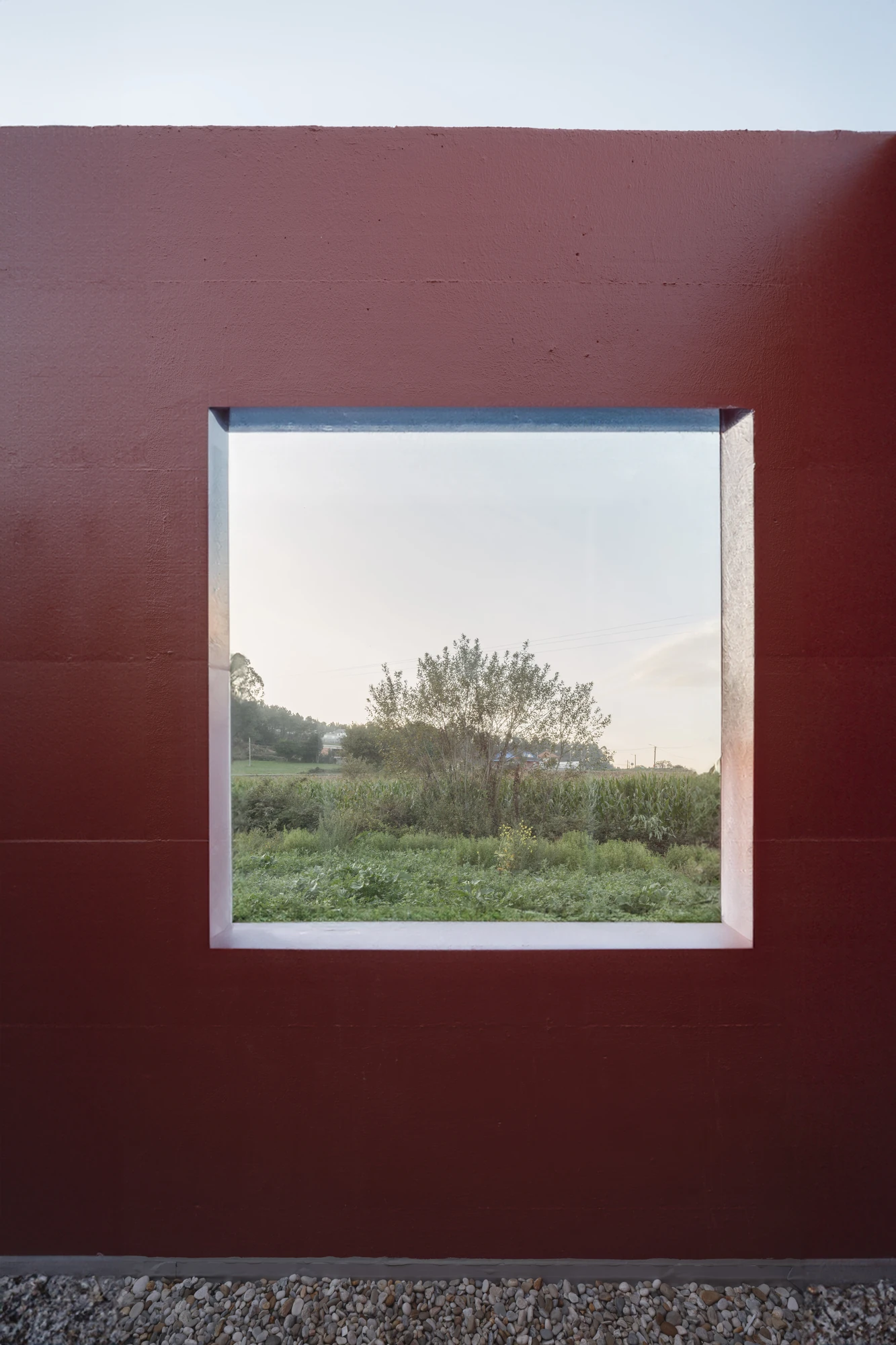 Goians Community Center by NULA.STUDIO + FCA, photographed by Luis Díaz Díaz / NULA.STUDIO — A deep square window punched through the oxide-red wall frames corn, trees and a distant village over a bed of gravel.