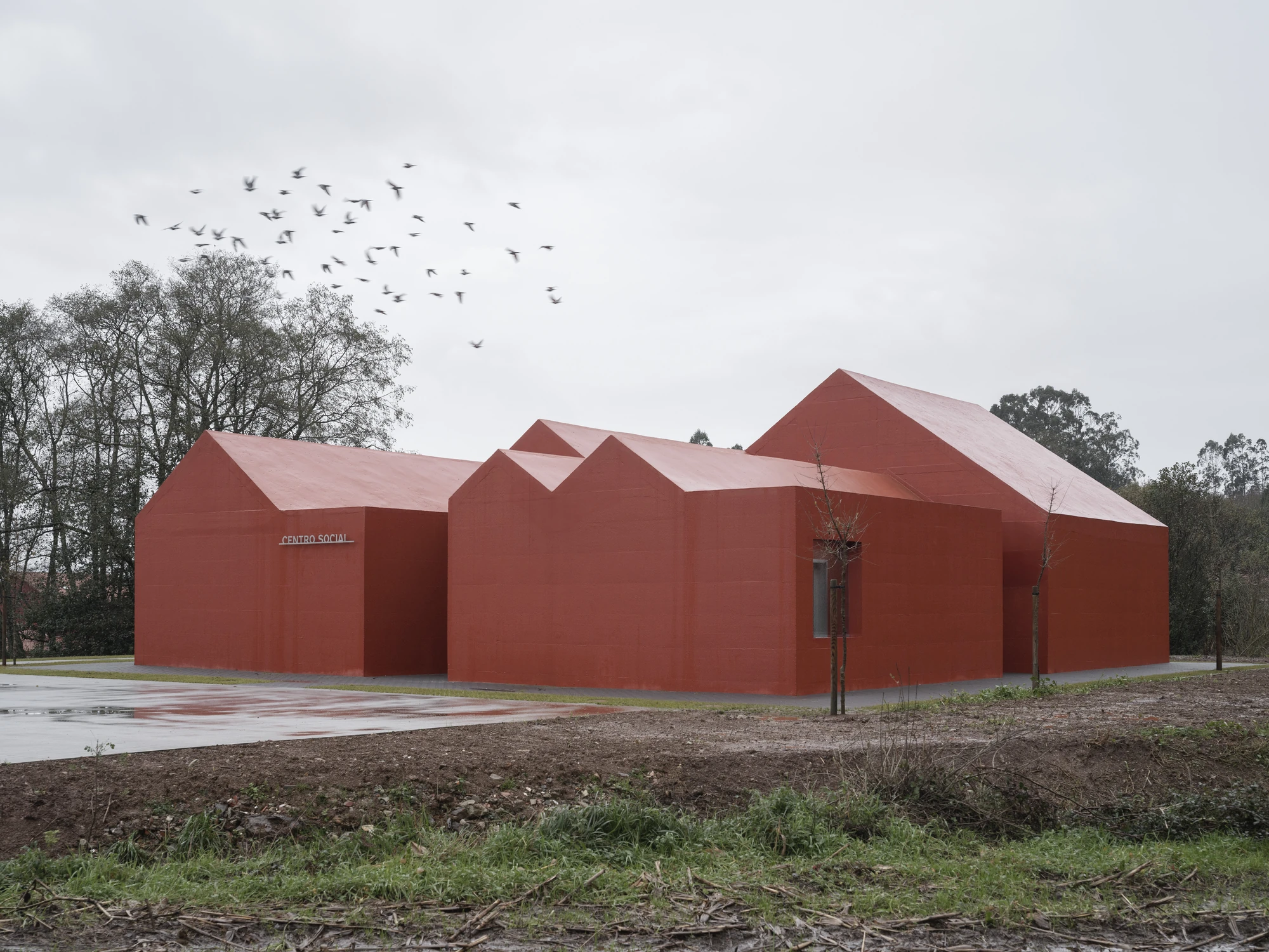 Goians Community Center by NULA.STUDIO + FCA, photographed by Luis Díaz Díaz / NULA.STUDIO — Under an overcast sky a flock of birds crosses above the red gabled mass, its wet concrete apron reflecting the grey day.