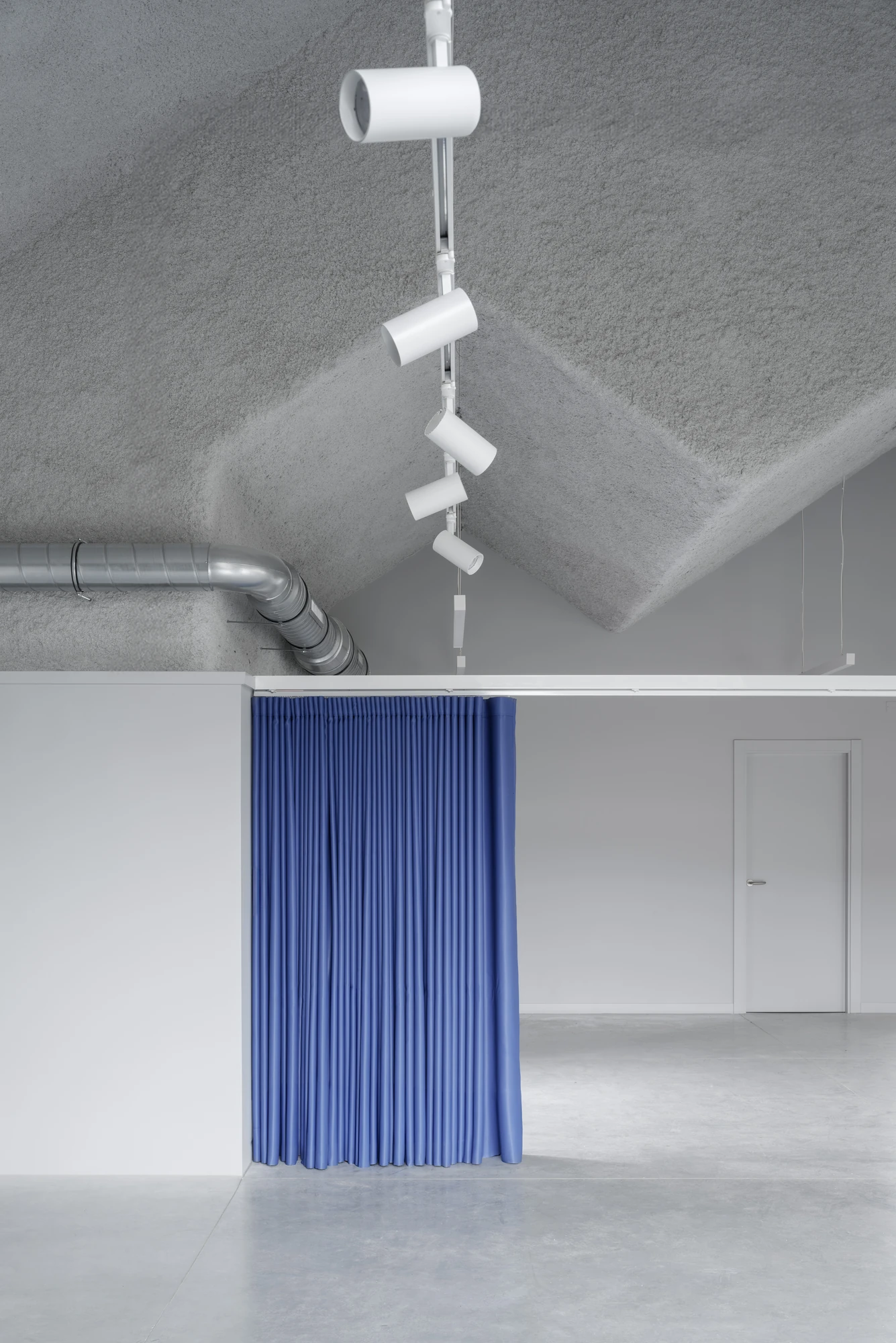 Goians Community Center by NULA.STUDIO + FCA, photographed by Luis Díaz Díaz / NULA.STUDIO — The folded grey ceiling drops toward a blue curtain, a run of white track spotlights and exposed ductwork threading the peak.