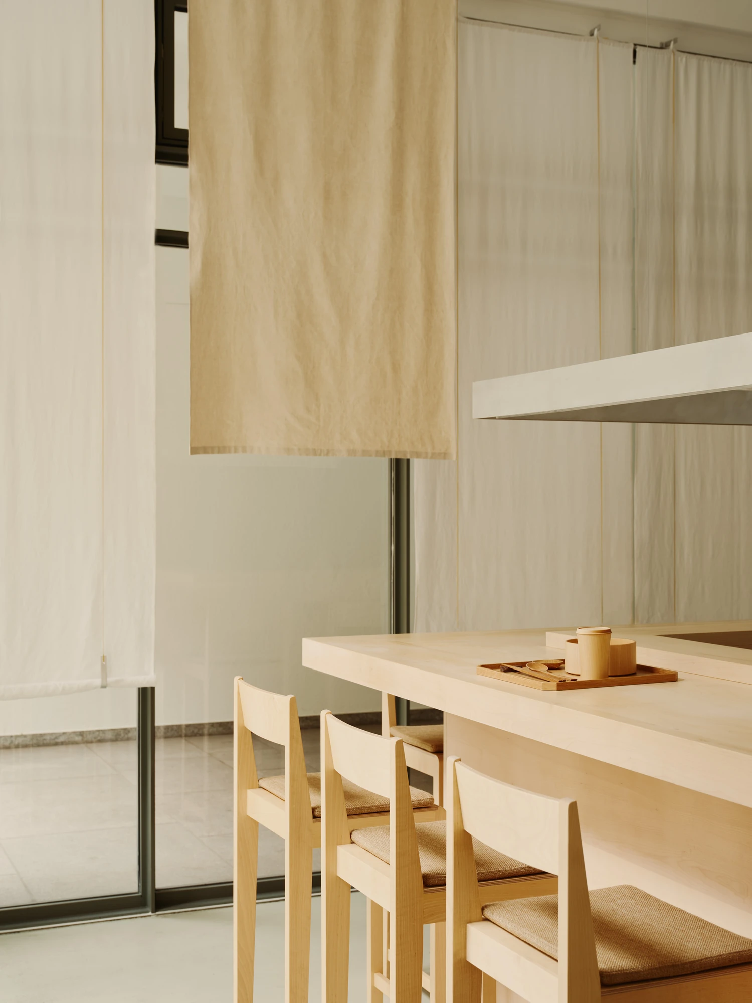 Nichigetsu by Studio Inhalt_, photographed by Robert Rieger — Hanging beige linen curtains filter daylight behind the maple counter, where a wooden tray holds turned cups and a spoon beside pale timber chairs.