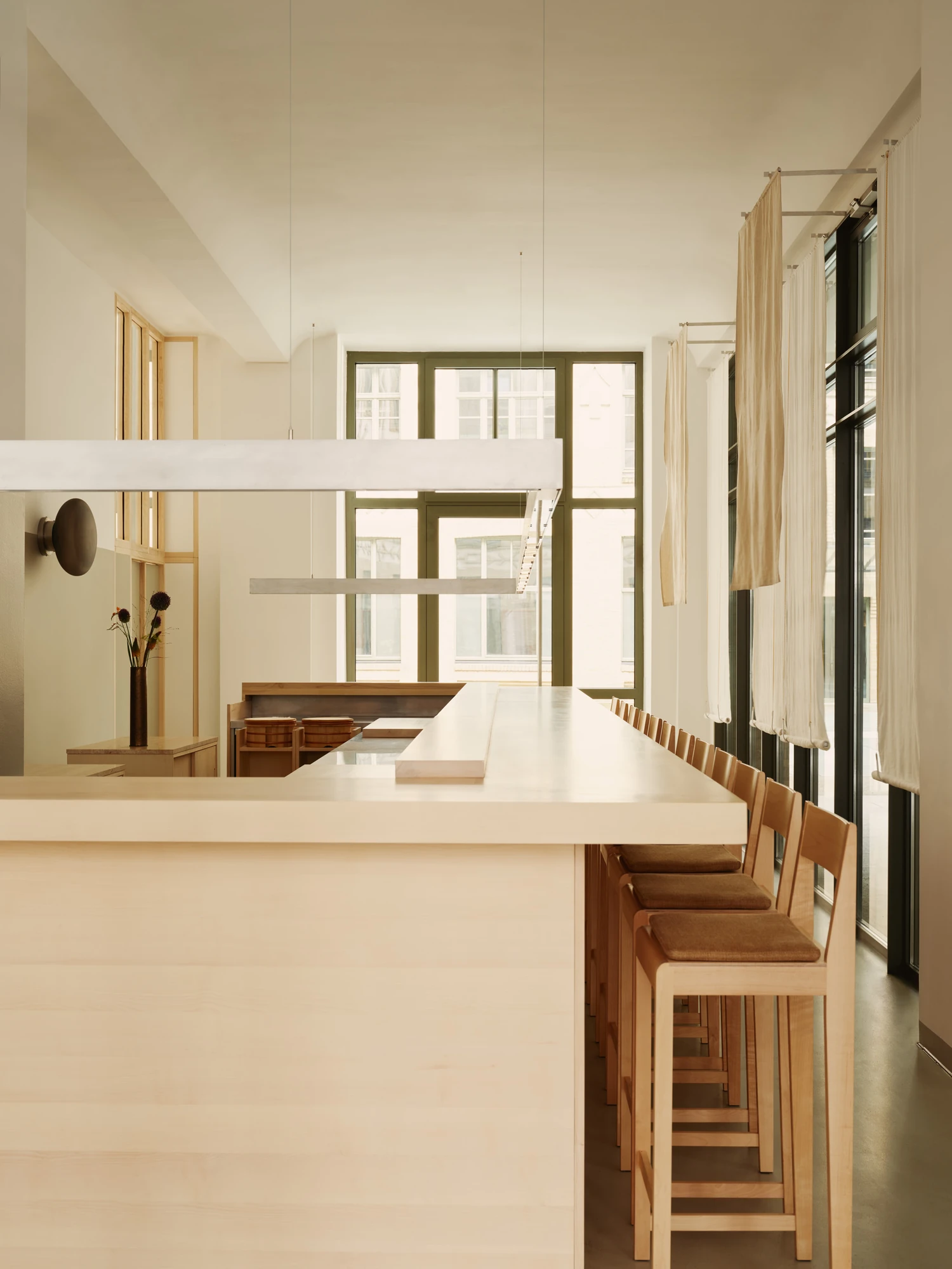 Nichigetsu by Studio Inhalt_, photographed by Robert Rieger — The long maple counter stretches toward tall green-framed windows, a steel pendant bar overhead and cedar rice tubs set into the chef's station.