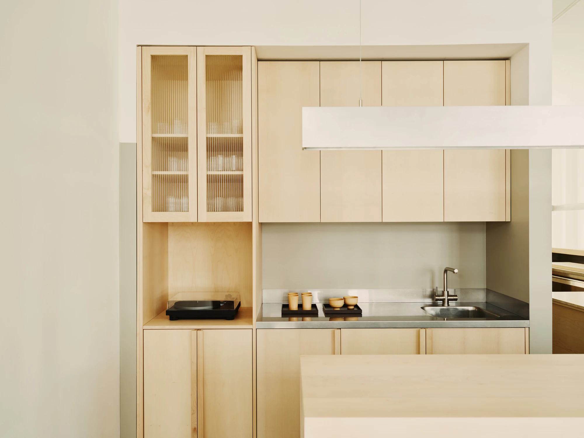 Nichigetsu by Studio Inhalt_, photographed by Robert Rieger — Light maple kitchen fronts and a reeded-glass cabinet frame a stainless-steel worktop, a record player nested in an open niche below the wall units.