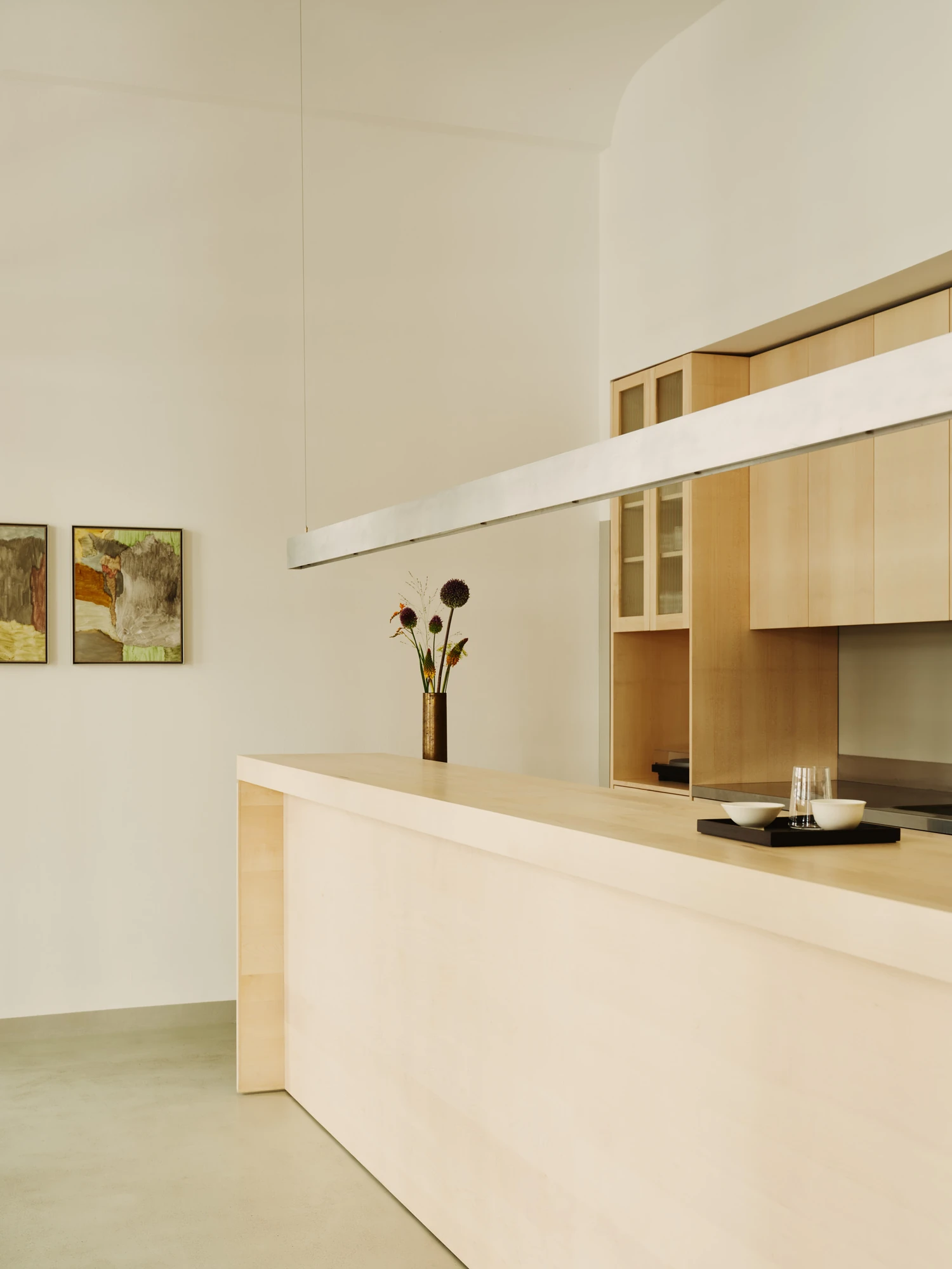 Nichigetsu by Studio Inhalt_, photographed by Robert Rieger — Two framed abstract paintings hang beside the maple counter under a linear steel pendant, an ikebana stem rising from a dark bronze vase.