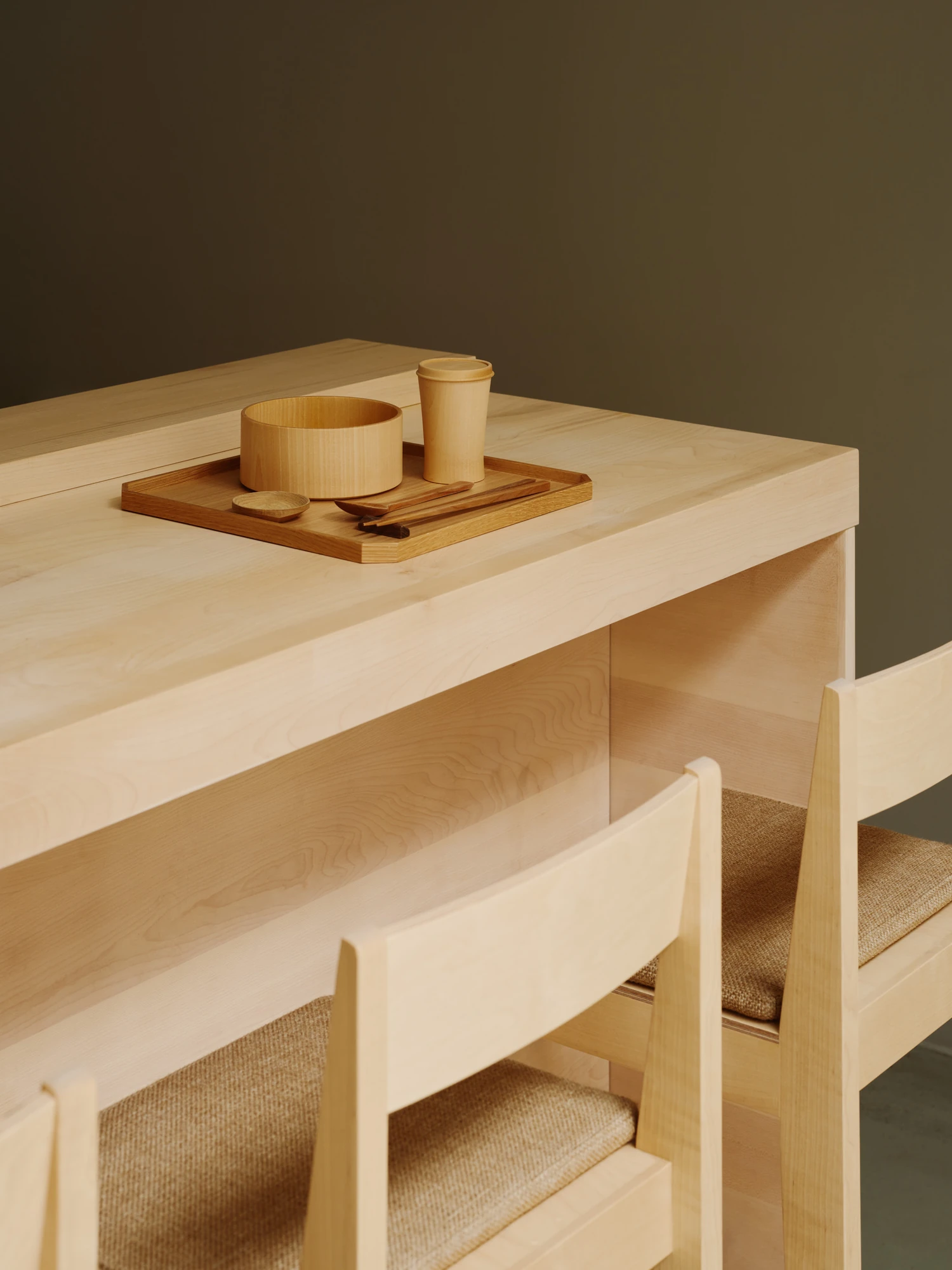 Nichigetsu by Studio Inhalt_, photographed by Robert Rieger — A corner of the maple counter, its edge thick and softly rounded, carries a wooden tray of turned vessels above chairs with woven seats against an olive wall.